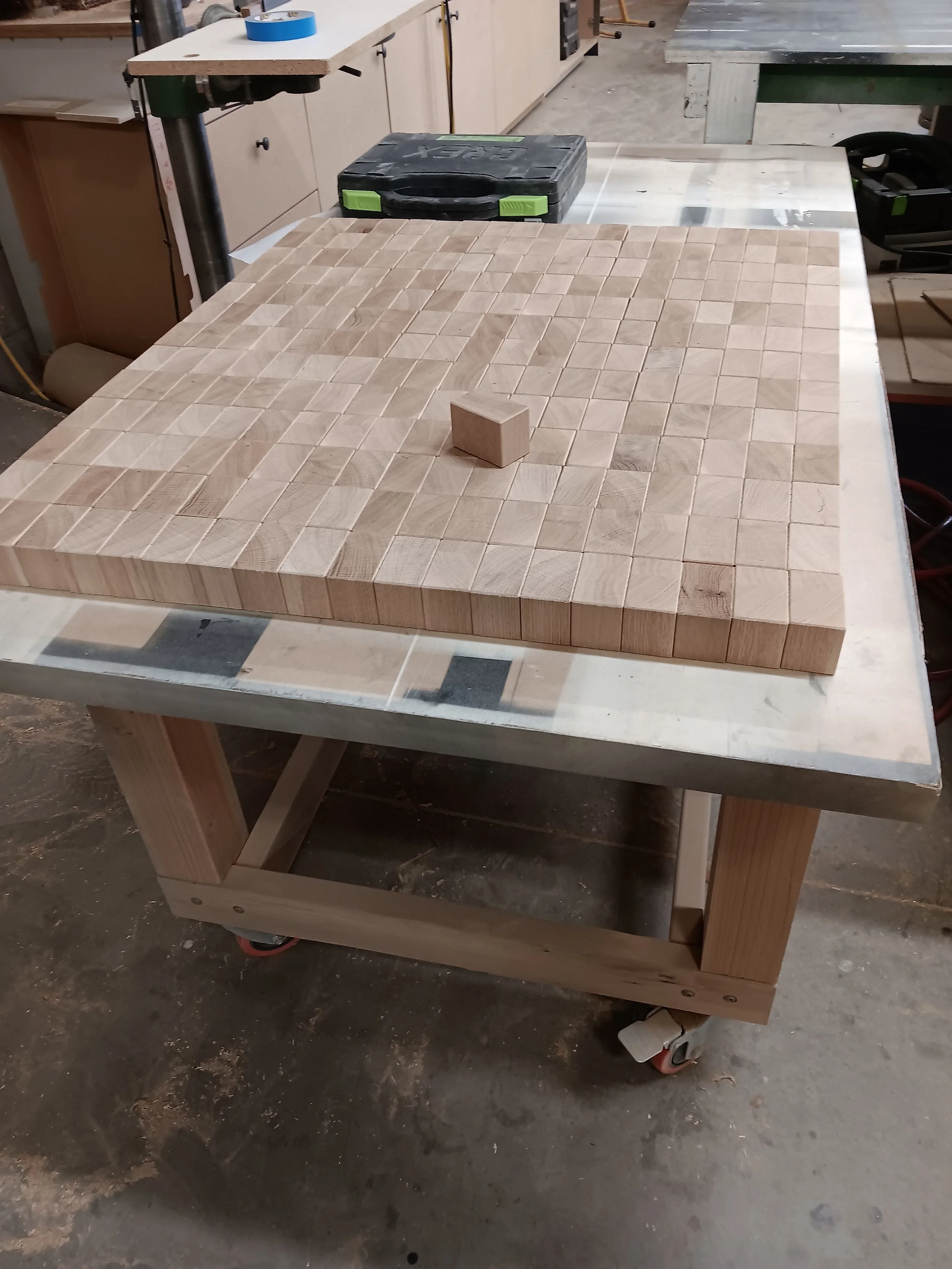 A woodworking worktable with a large square wooden mosaic grid and a small wooden cube on top, in a carpentry workshop.