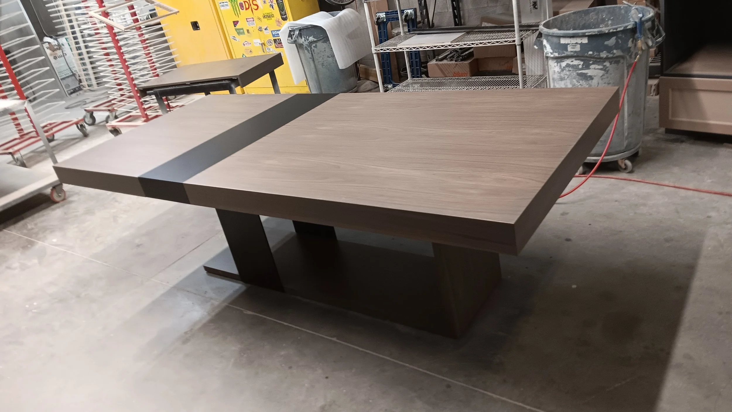 A wooden rectangular dining table with a dark base in a warehouse or storage area surrounded by carts, shelves, a trash bin, and yellow vending machines in the background.