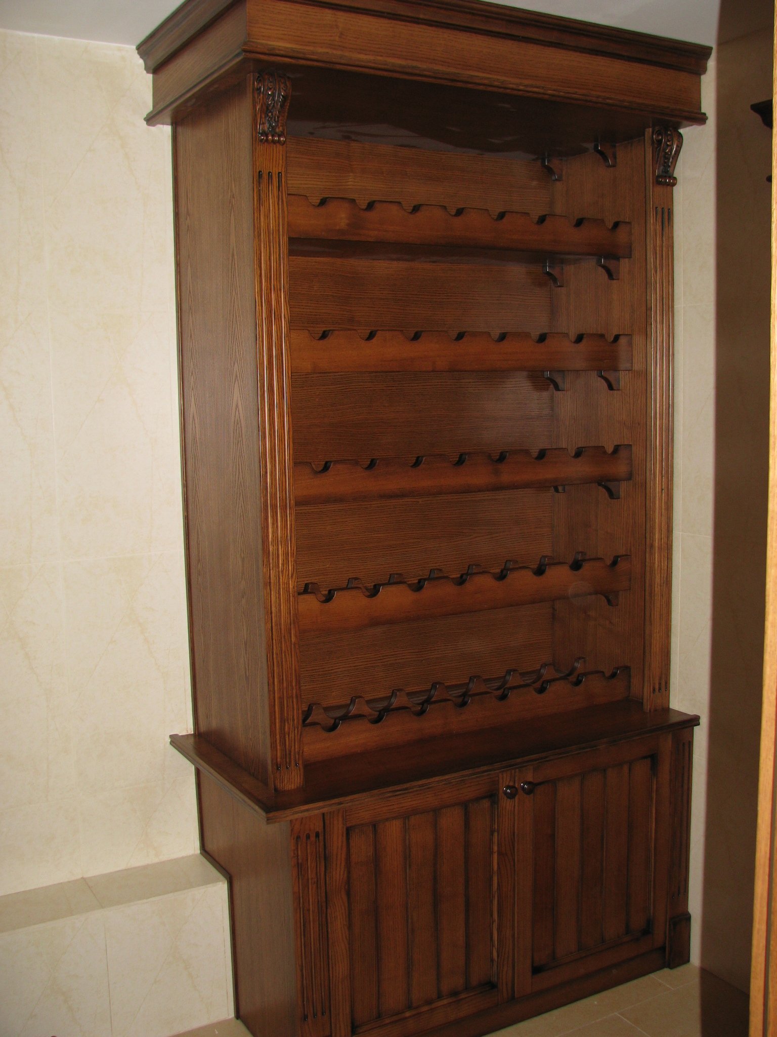 Interior view of a modern home with a wooden corner cabinet and a glass display case containing decorative items and bottles, next to a door with horizontal black stripes, and a dining area in the background with a white table and chairs near a window.