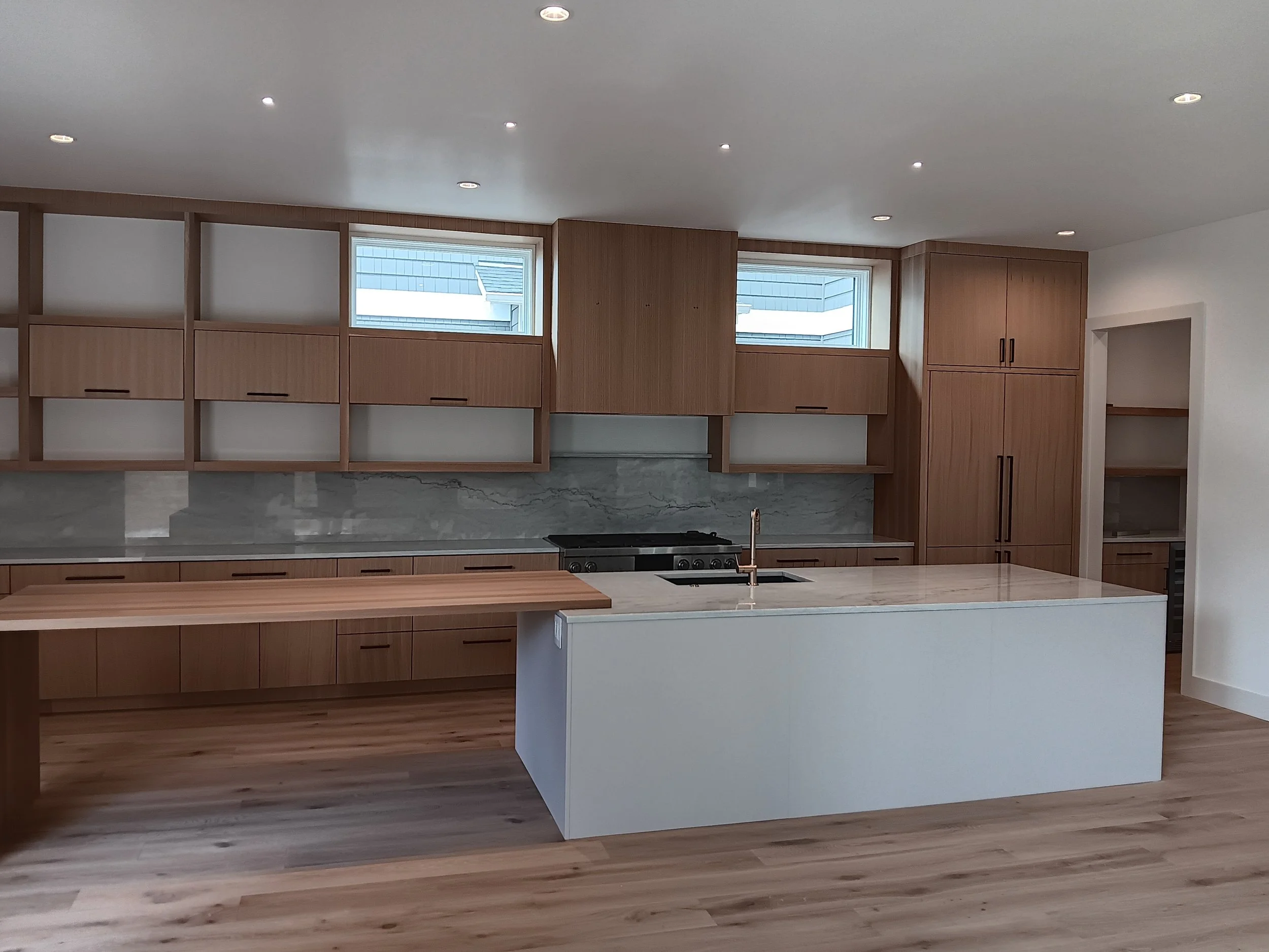 Modern kitchen with wooden cabinets, a kitchen island with a marble countertop, and a gray marble backsplash. Two small windows are on the wall above the cabinets. Recessed ceiling lights illuminate the space.