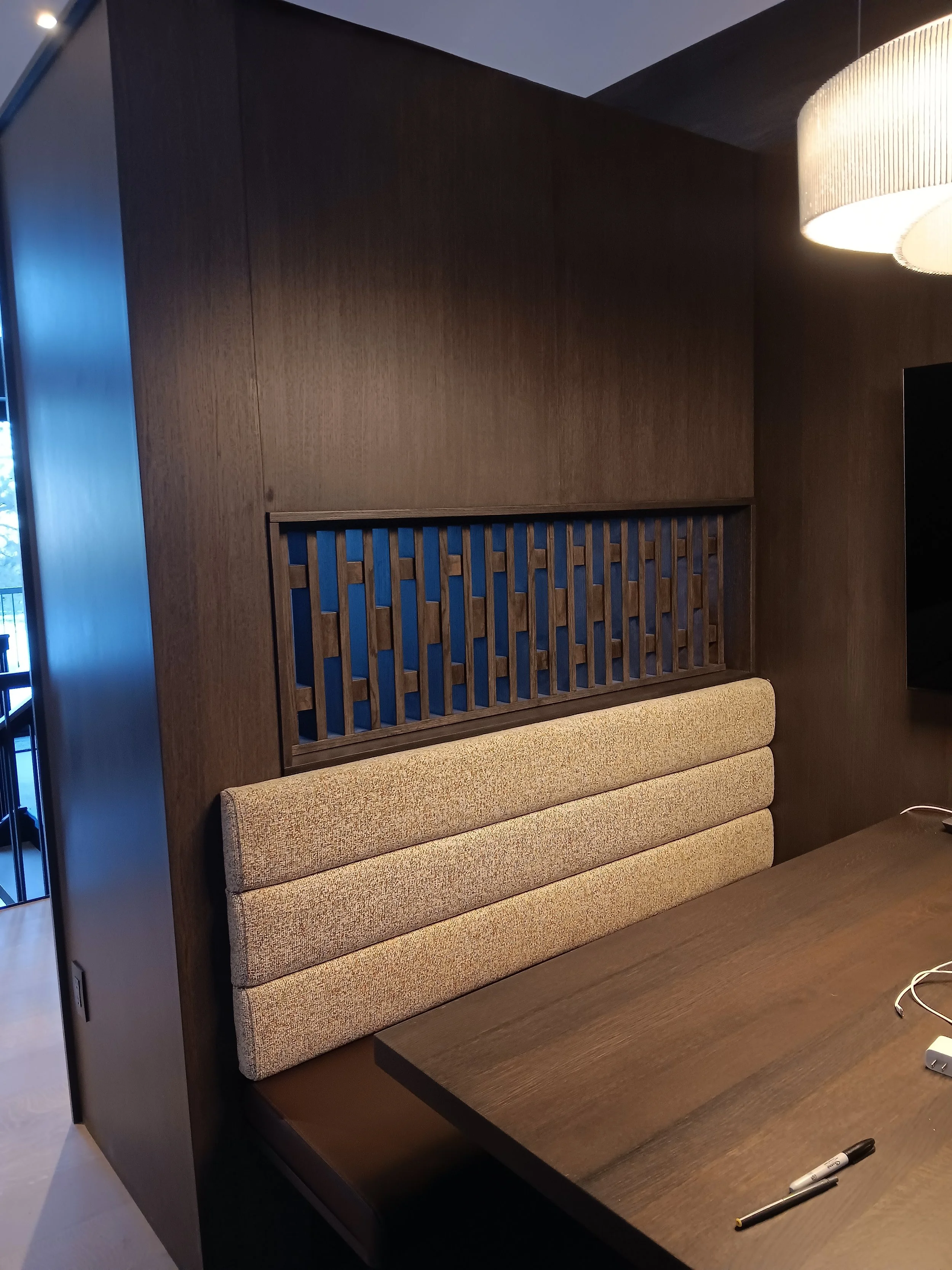 Wood-paneled wall with a decorative wood lattice screen above a beige cushioned booth seat, next to a dark wooden table with a black marker and some electronic cables on it.