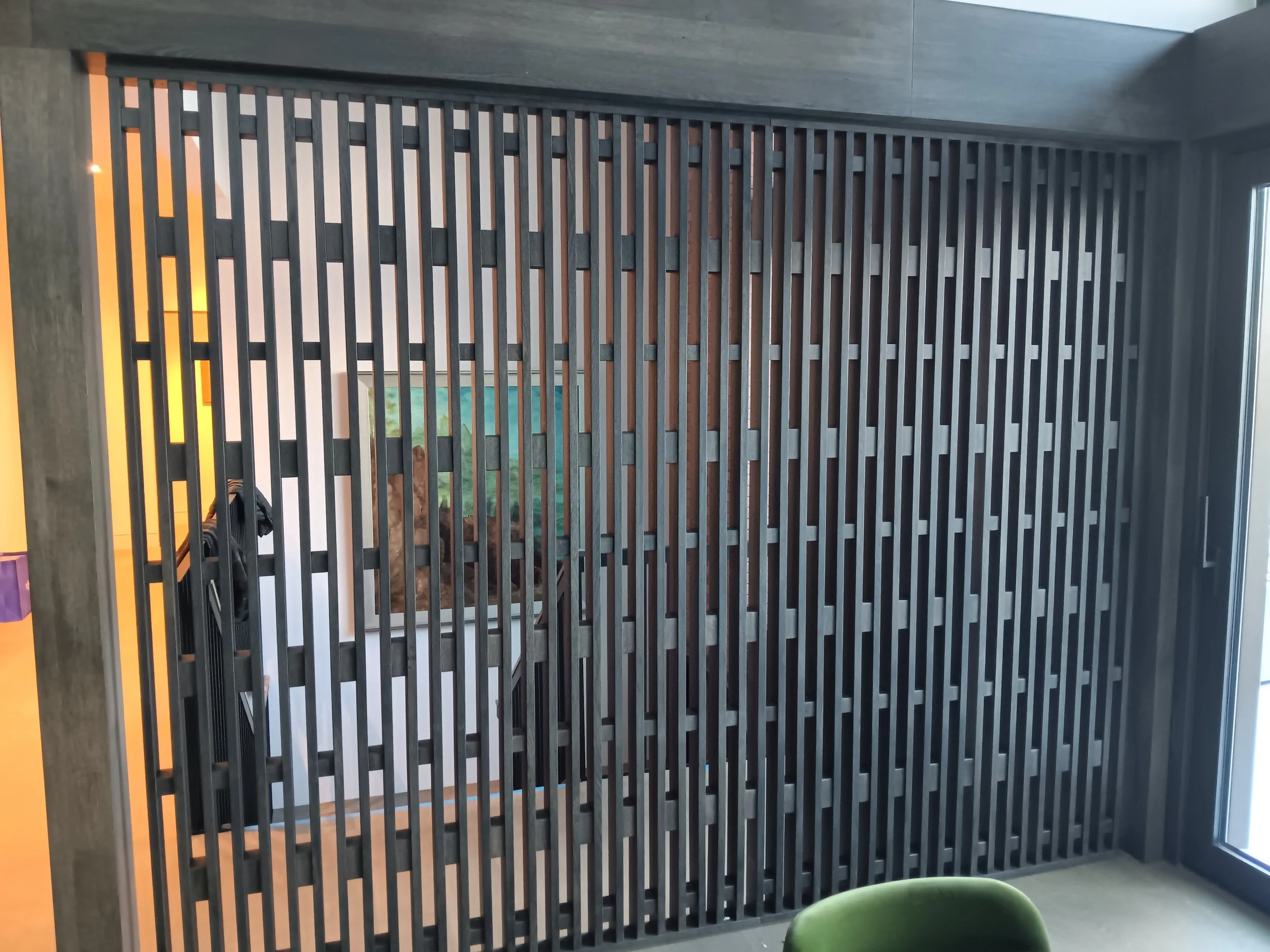 Modern interior with a vertical wooden slat partition wall, a window on the right, and a glimpse of a room with artwork and furniture behind the slats.