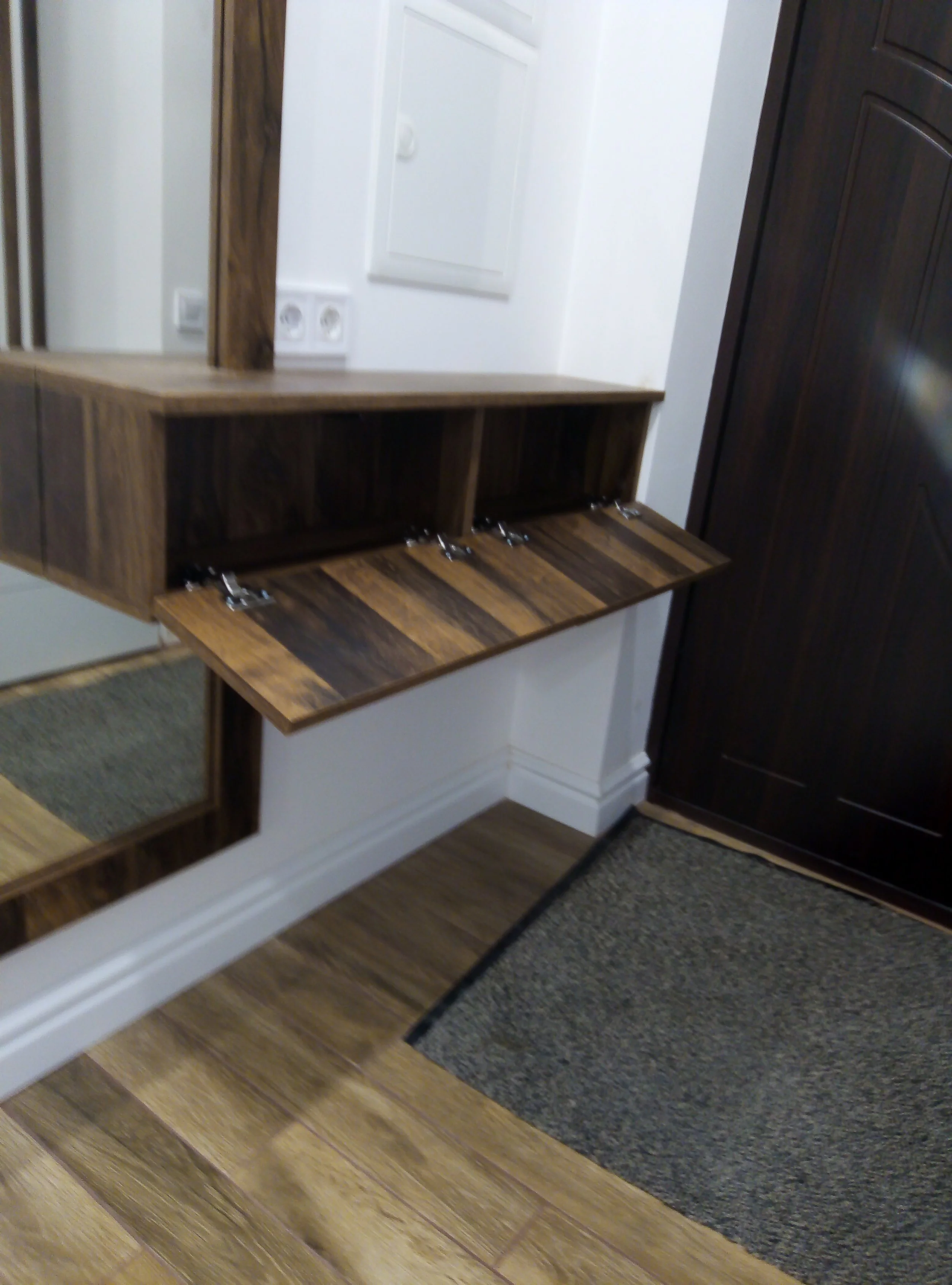 A wooden wall-mounted foldable shelf with compartments, next to a mirror, a dark wooden cabinet, and a gray rug on a wooden floor.