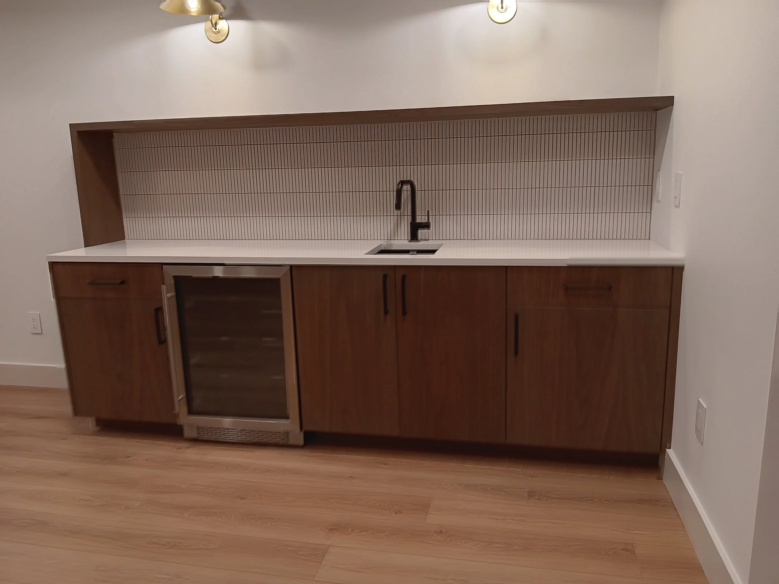 Modern kitchenette with wooden cabinets, a white countertop, a small undermount sink with a black faucet, a built-in wine fridge, and vertical striped backsplash tiles, under two wall-mounted light fixtures.