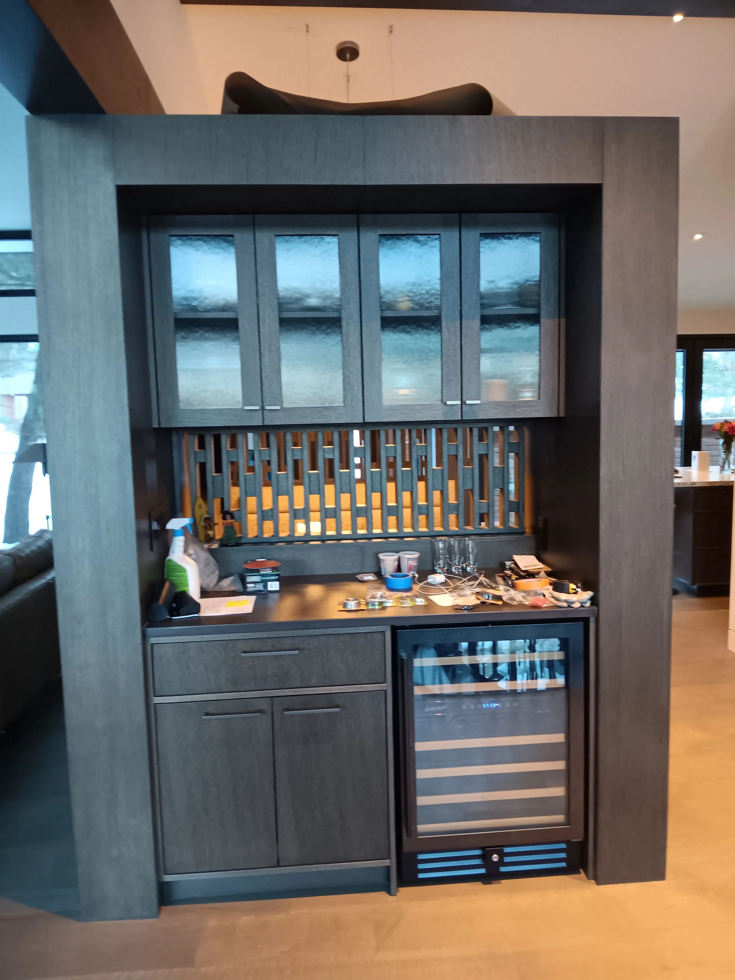 A small home bar area with cabinets, a wine cooler, and various bottles and glasses on the countertop.