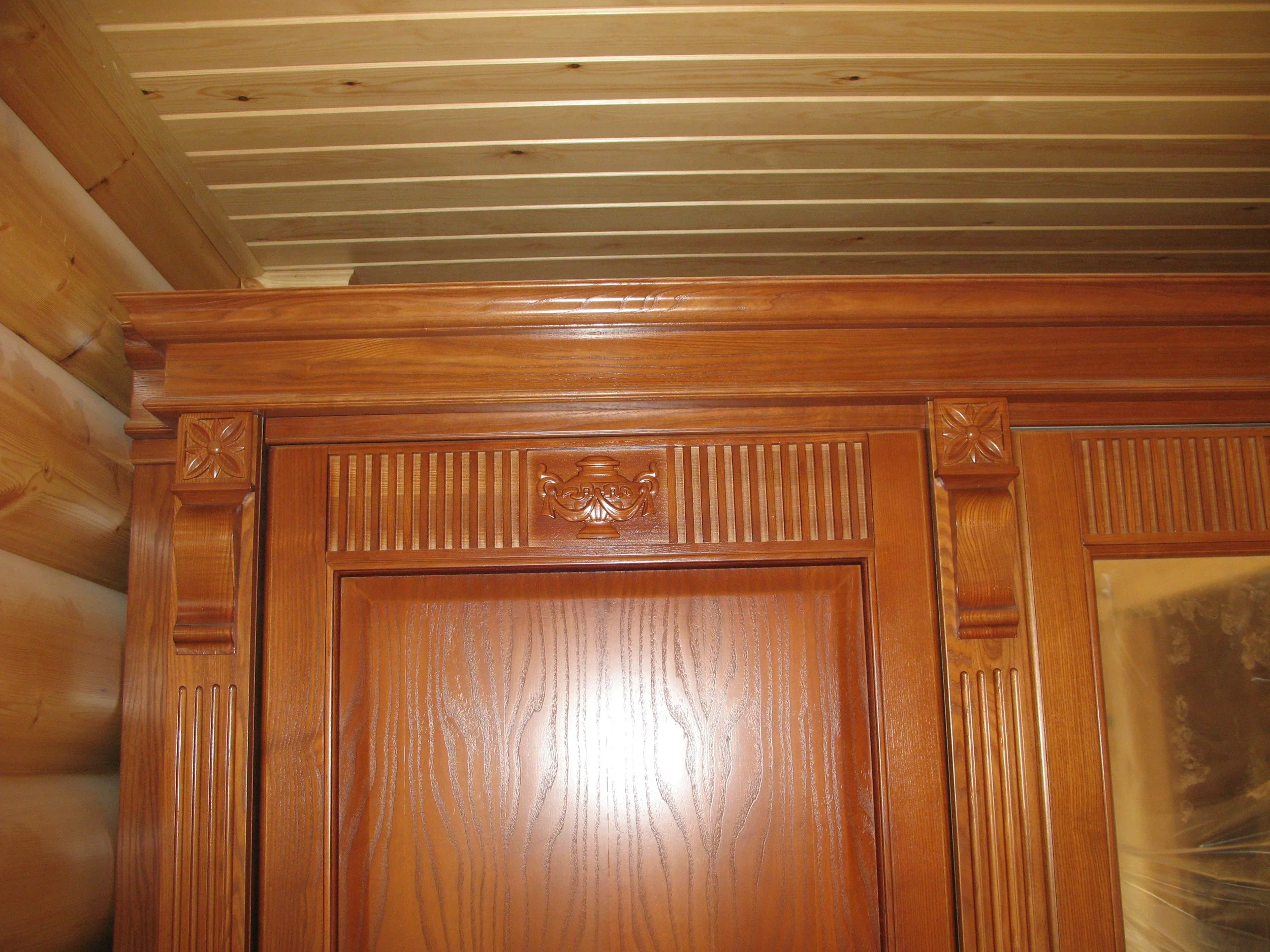 Close-up of a wooden doorway or cabinet with decorative carving and molding. The wood has a polished finish, showcasing the grain patterns.