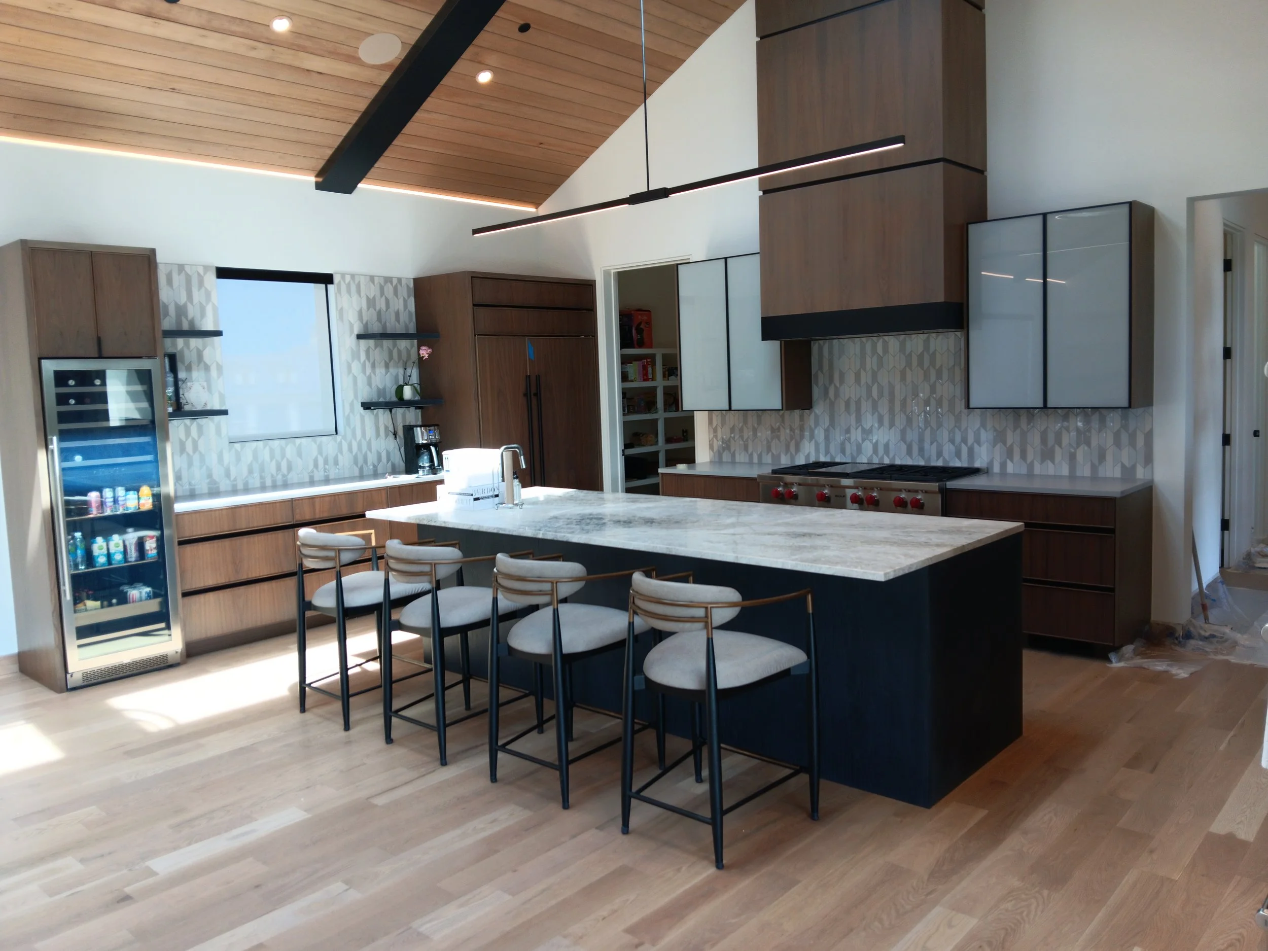 Modern kitchen with a large island, wooden cabinets, gray and white patterned backsplash, bar stools, and a glass-front wine cooler.