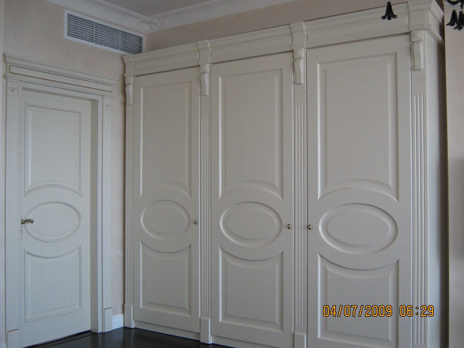 A corner of a room with white decorative built-in wardrobes and a door with matching design, along with a wall vent above the door.