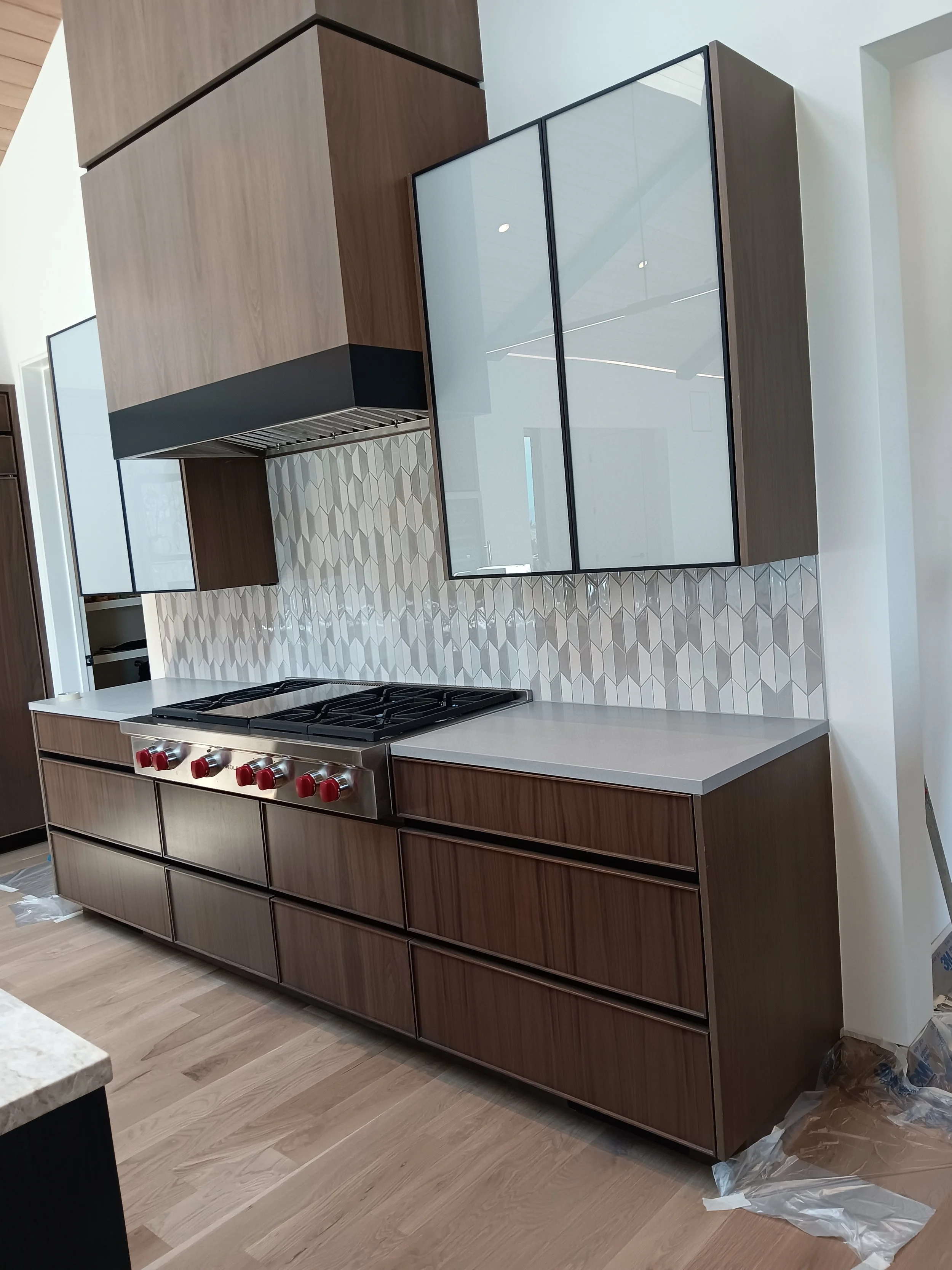 Modern kitchen with dark wood cabinets, a range with red knobs, light gray countertops, a geometric tile backsplash, and glass-front upper cabinets.