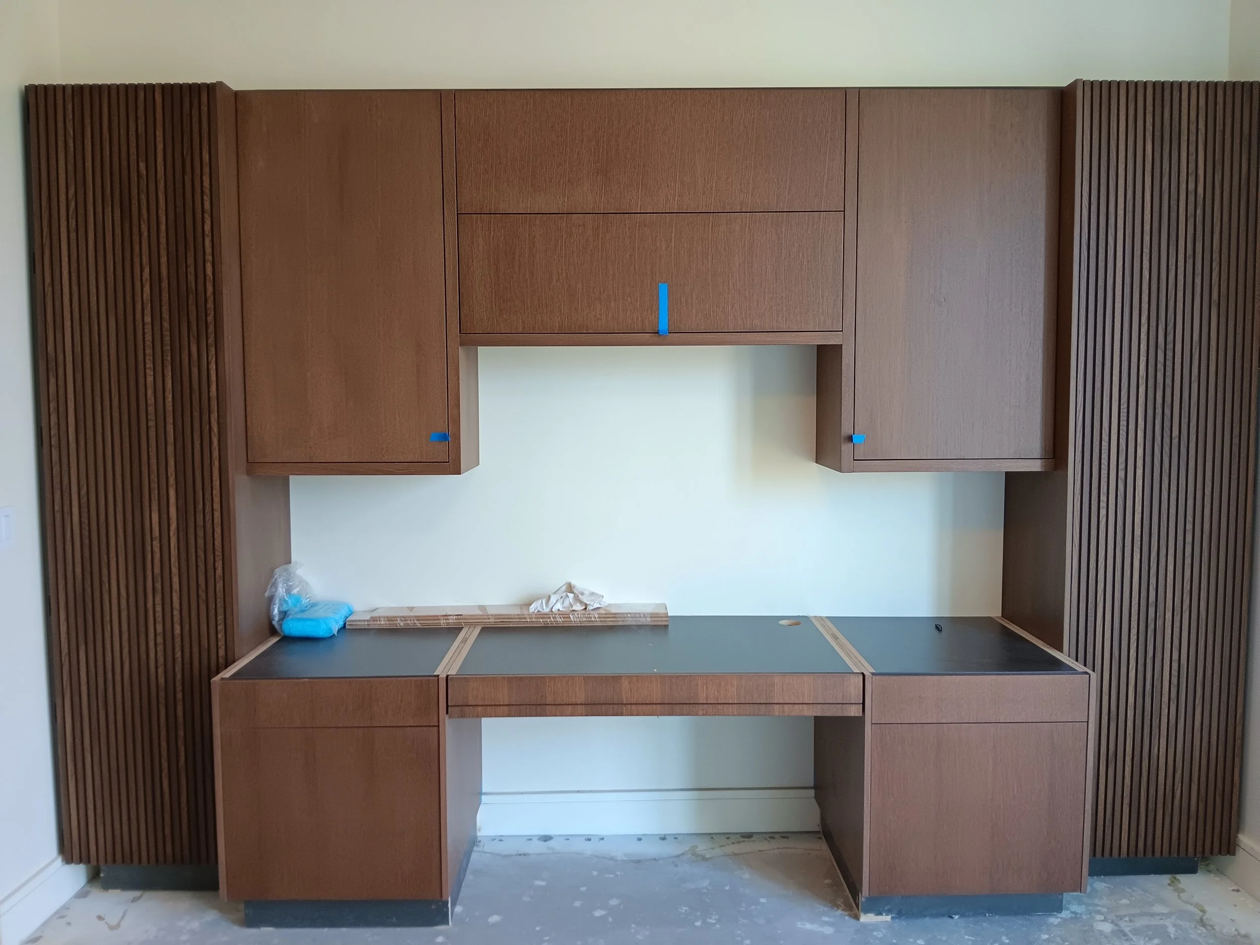 Installation of wooden cabinets in a kitchen or office space, with some parts still unfinished and blue painter's tape on the cabinets.