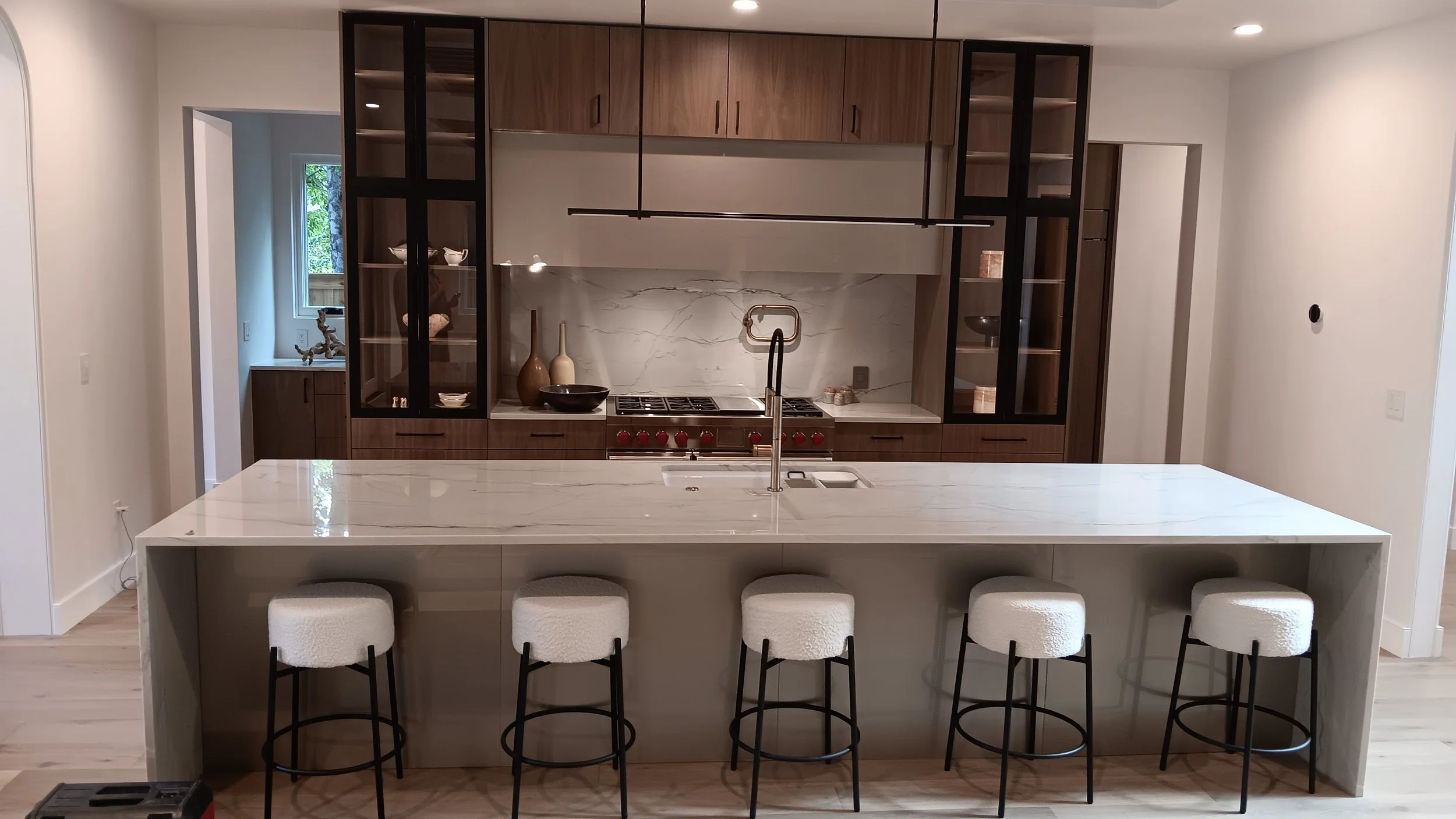 Modern kitchen with a large island, white marble countertop, five bar stools with white cushions, wooden and glass cabinetry, a marble backsplash, and a stainless steel stove.