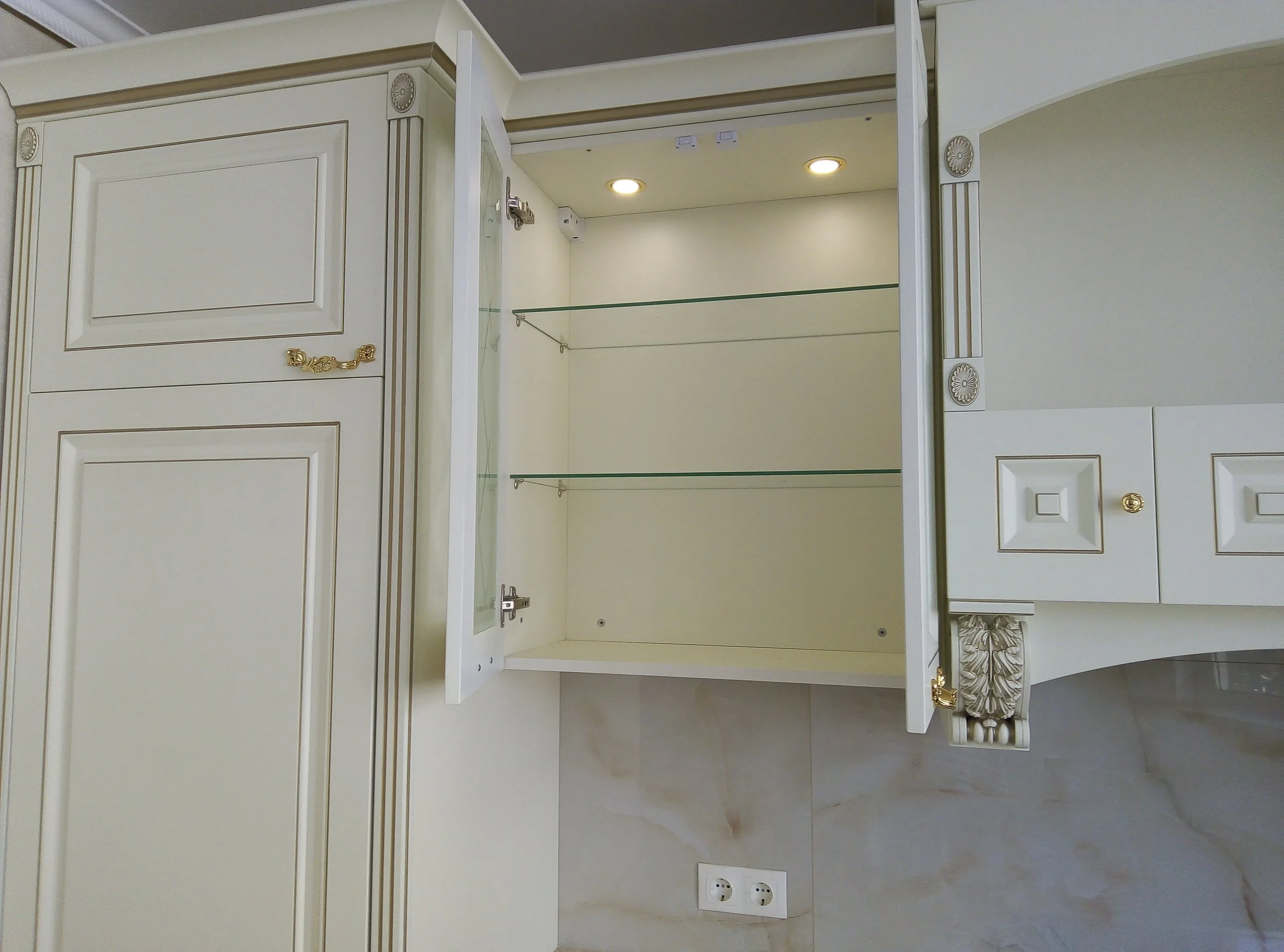 Empty white bathroom cabinet with glass shelves, ornate gold and wood details, and recessed lighting.