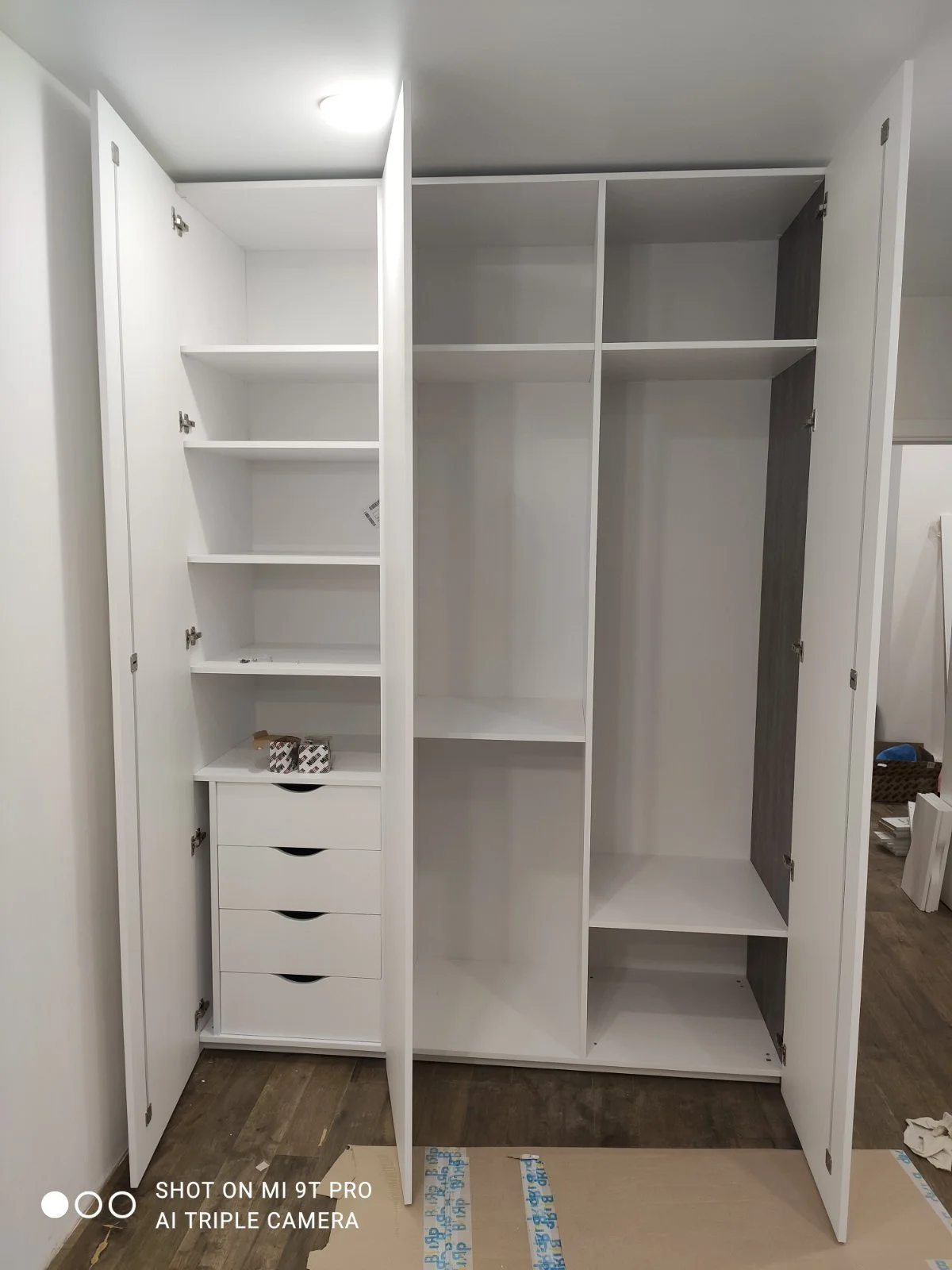 Empty white wardrobe with open doors showing multiple shelves and drawers inside, placed on a wooden floor.