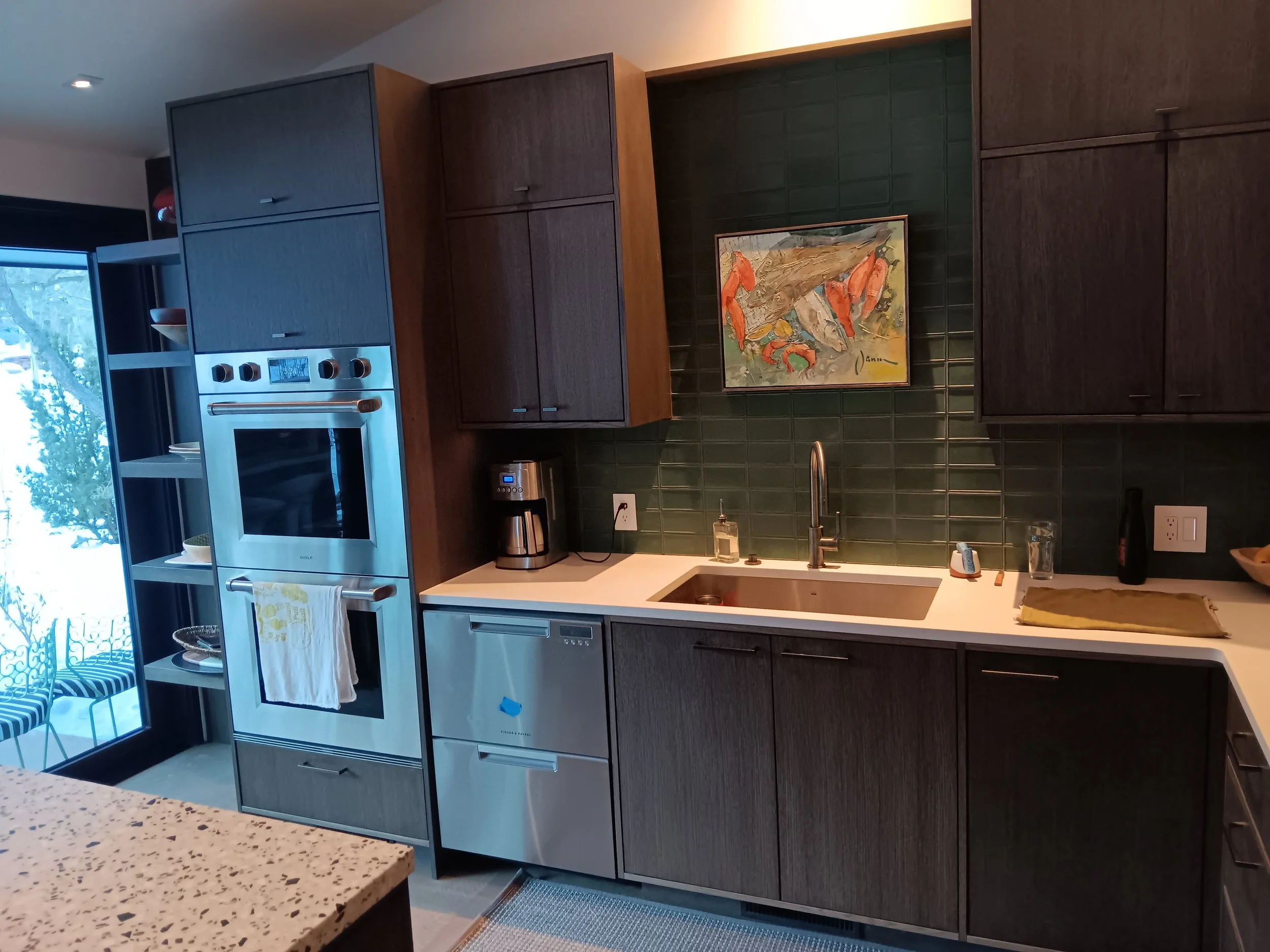 Modern kitchen with dark wood cabinets, green tiled backsplash, stainless steel oven and dishwasher, coffee maker, art painting of crabs, and a window with outdoor seating.