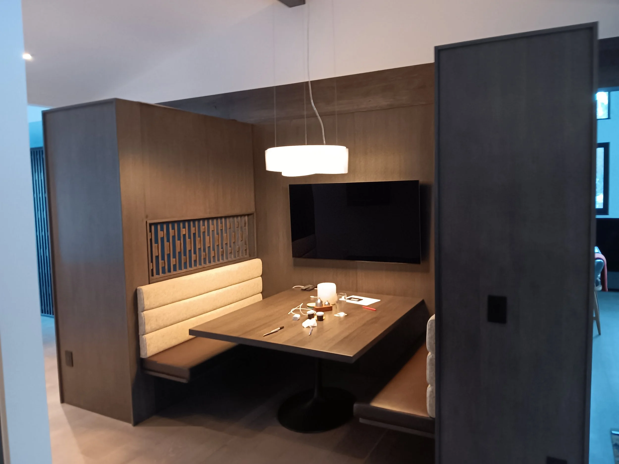 Modern dining nook with a wooden table, beige padded booth seating, a large flat-screen TV, a hanging light fixture, and various small objects on the table.