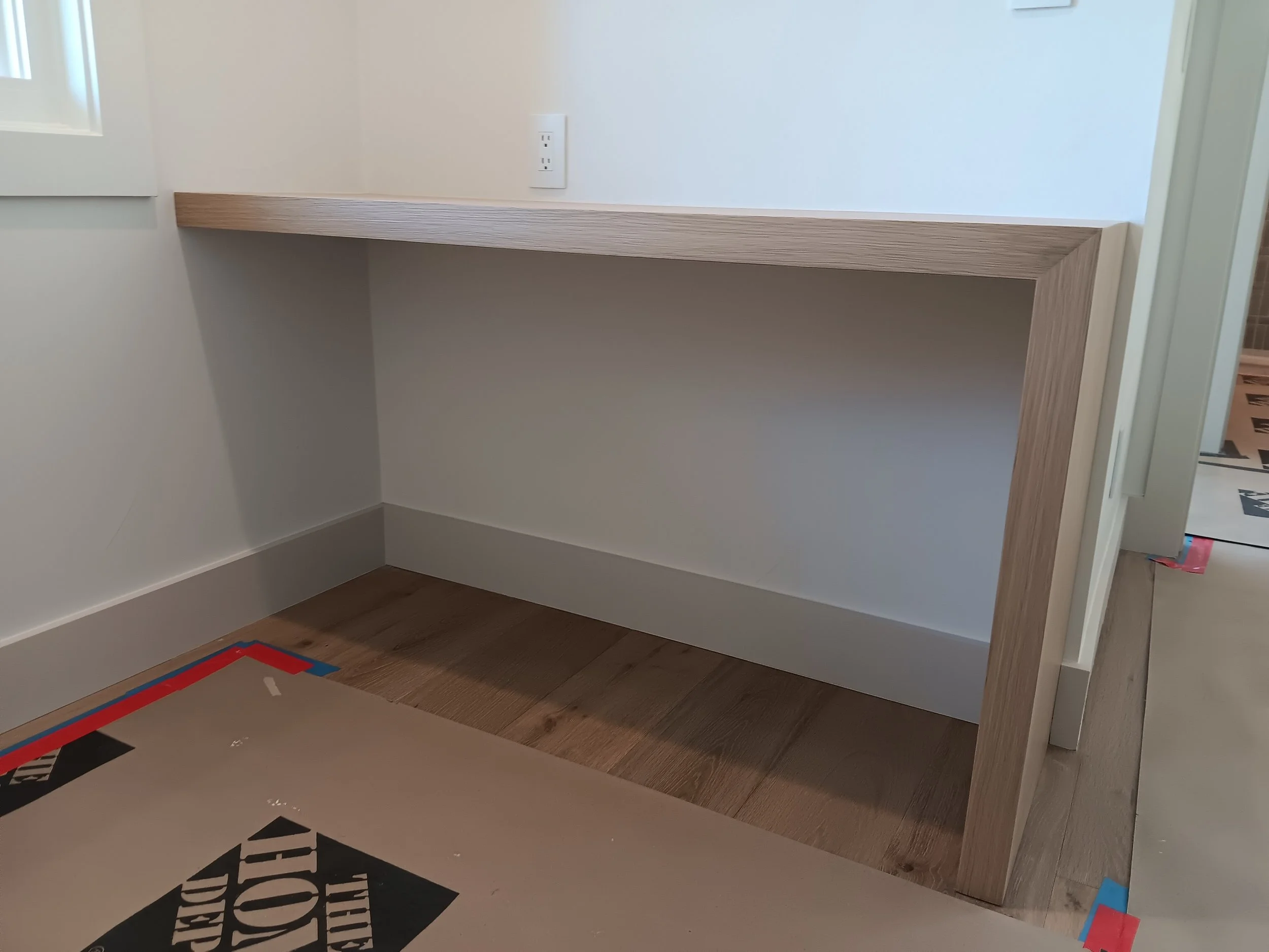 A partially built wooden desk or counter against a white wall in a room under construction, with a power outlet above and protective paper on the floor.