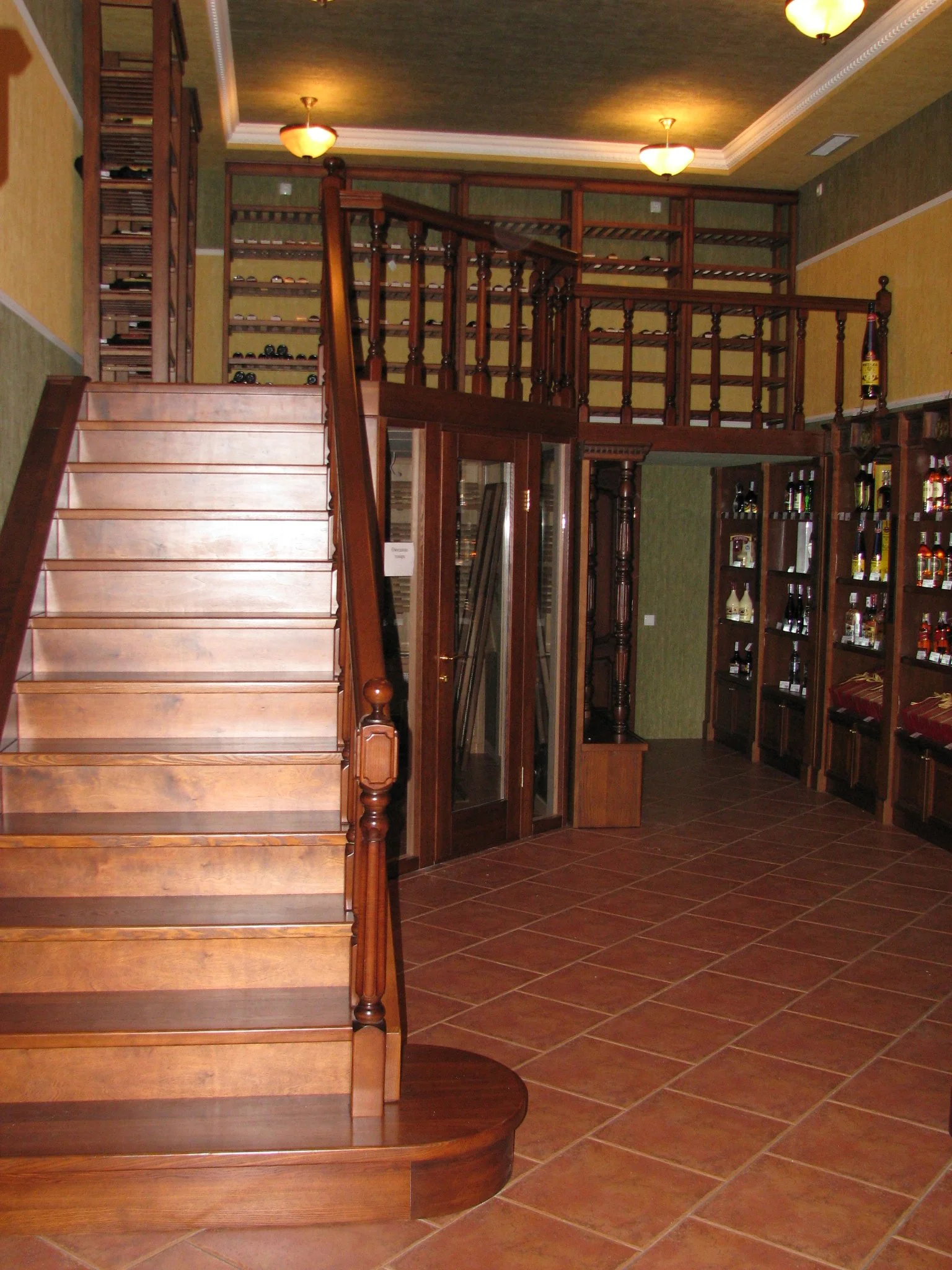 Interior of a wine cellar with wooden stairs leading to a second level, wooden shelves filled with wine bottles, and a tiled floor.