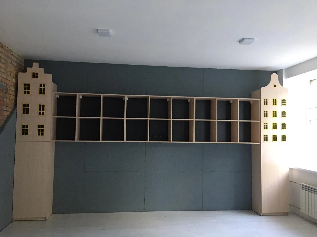 Empty wooden shelves with decorative house-shaped ends, black back panels, placed against a blue wall with a brick section on the left and a window on the right.