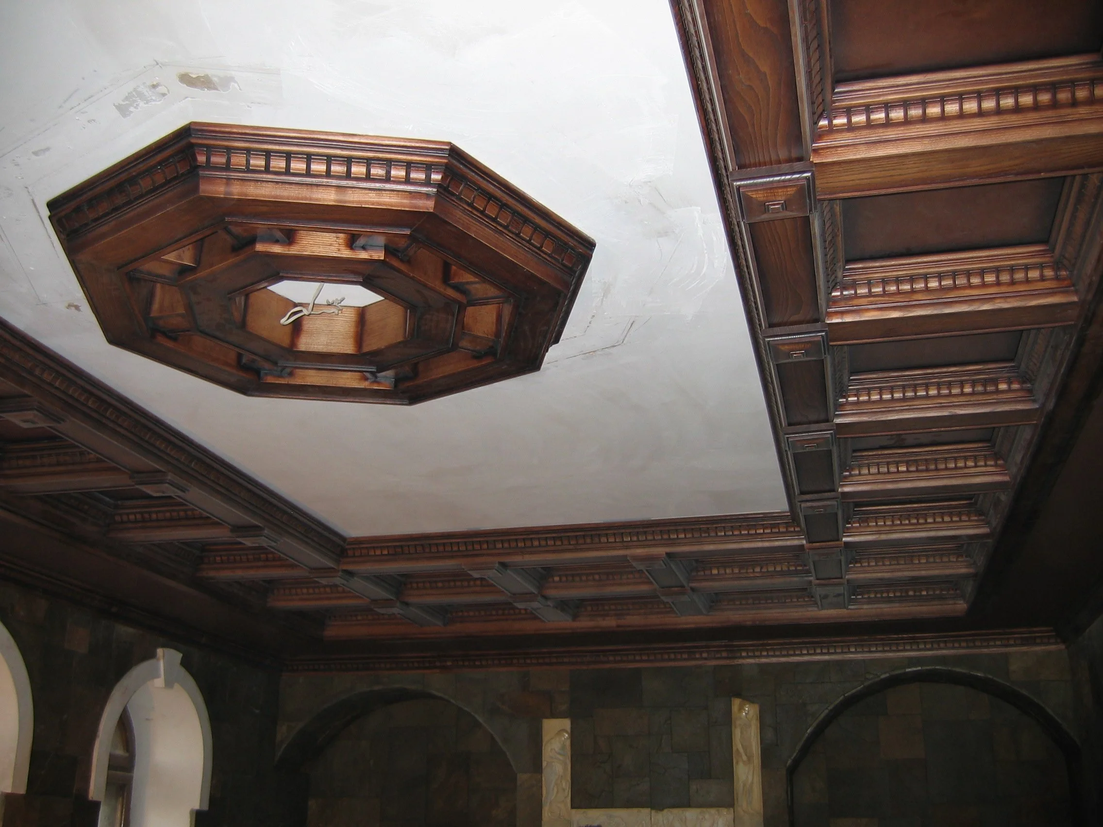 Decorative wooden ceiling panel with intricate molding and a central octagonal wooden fixture on a white ceiling.