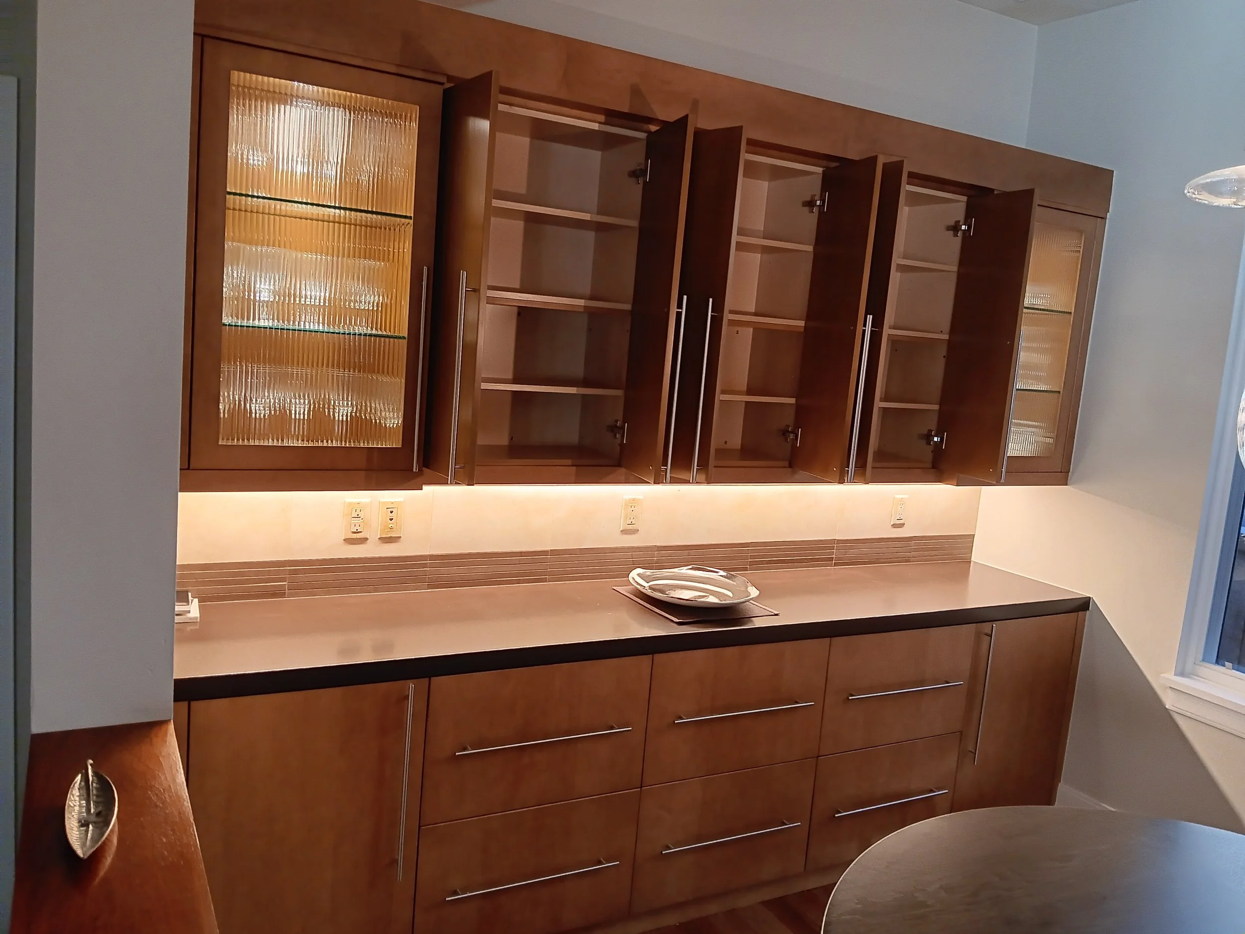 Empty kitchen cabinet with open upper glass-paneled doors and closed lower drawers and cabinets, under countertop with a plate and utensils.