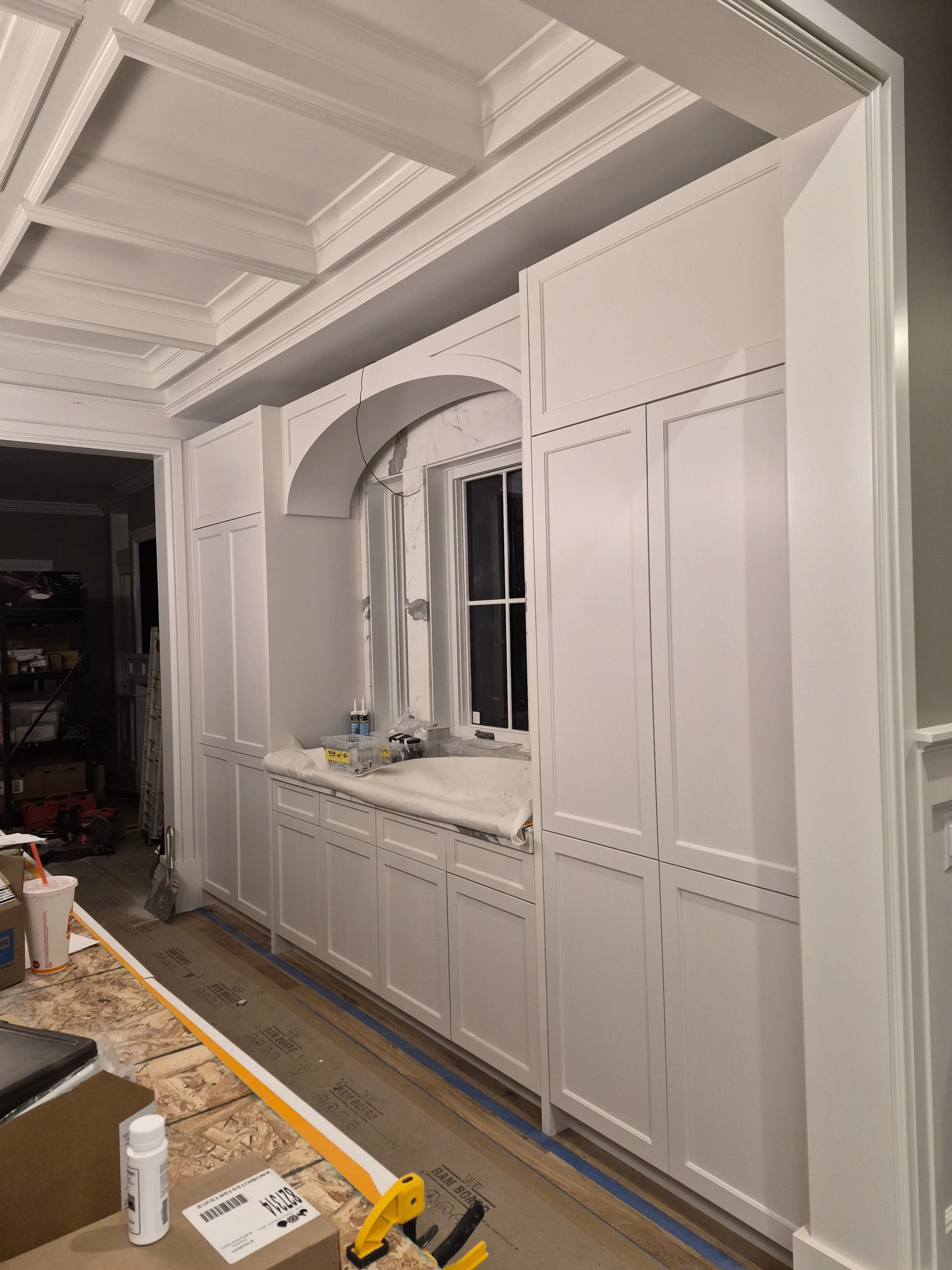 Interior view of a kitchen or pantry under construction, with white cabinetry, a window, and construction tools and materials on the floor.