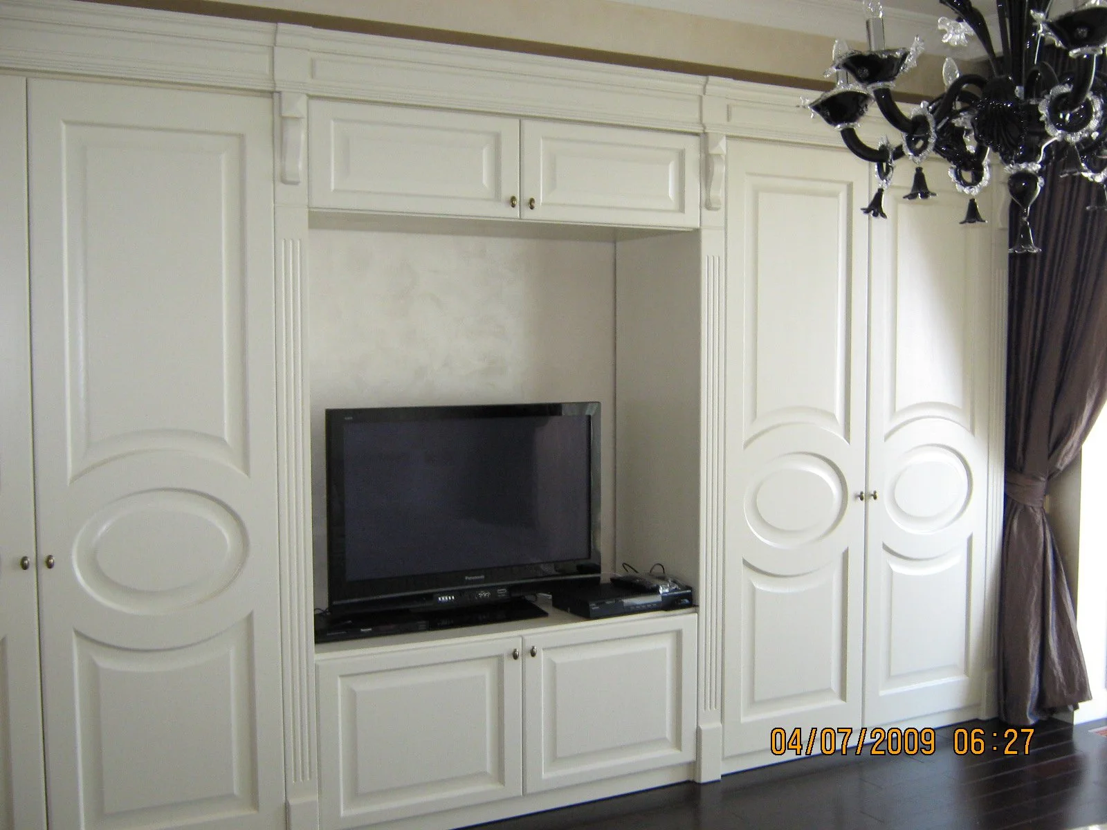 Living room entertainment center with white built-in cabinets, a black flat-screen TV, and a black chandelier with multiple arms hanging from the ceiling. There are dark curtains on the right side of the image.