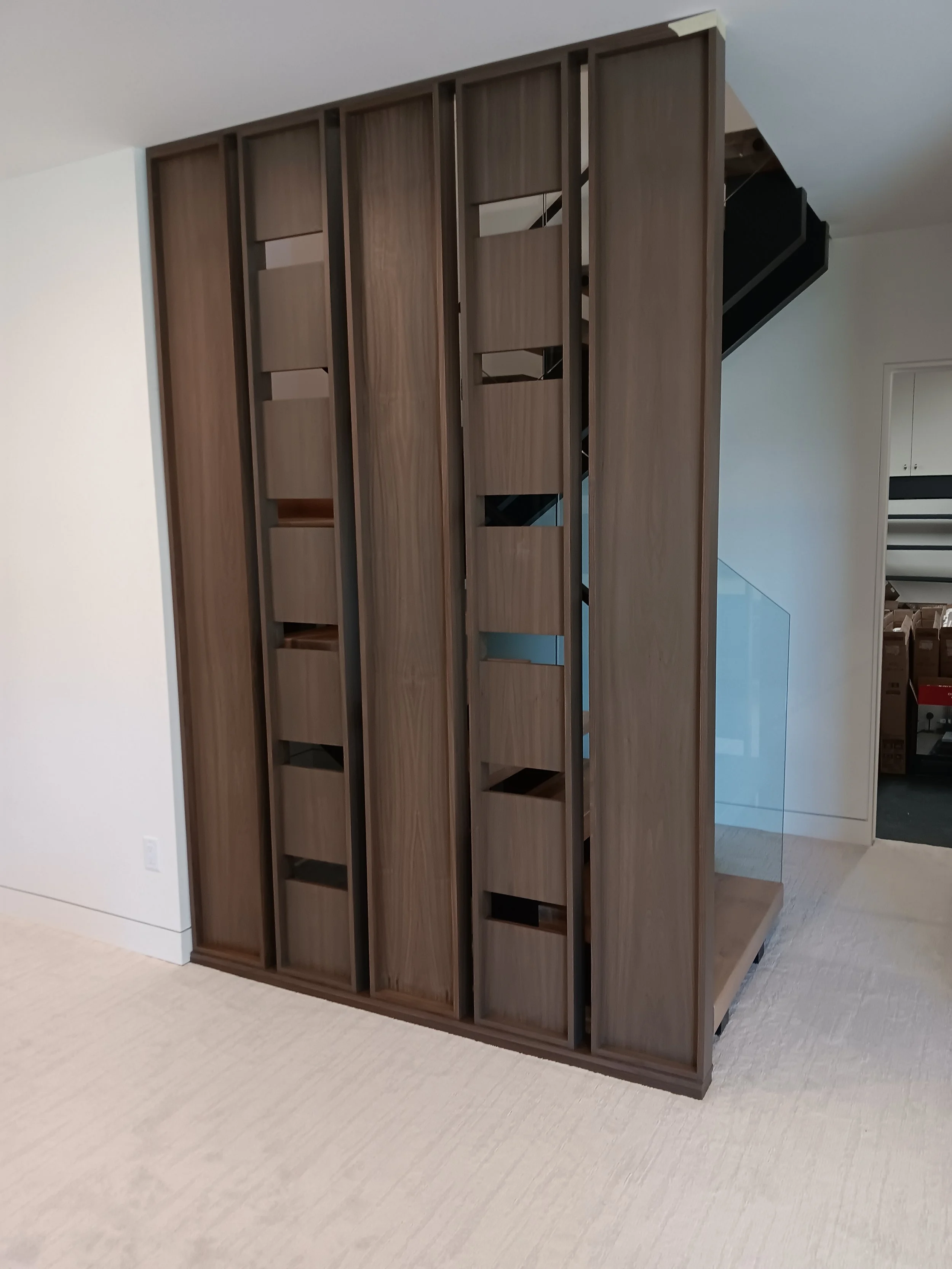 A wooden partition with vertical slats and open compartments, partially concealing a staircase with a glass railing in the background.