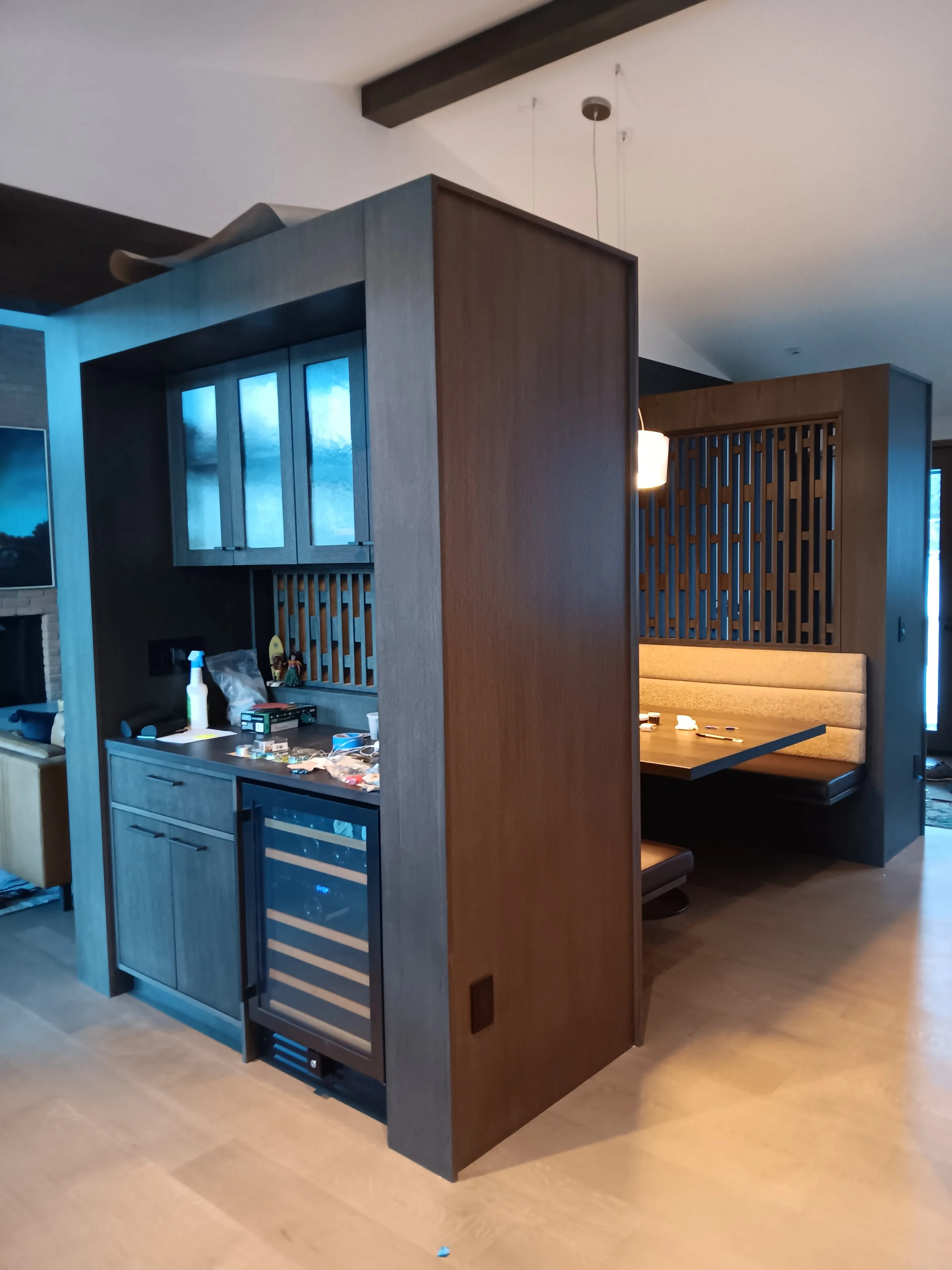 Interior view of a wooden partition with a built-in mini fridge and cabinets, separating a seating area with a booth-style dining nook inside a home.