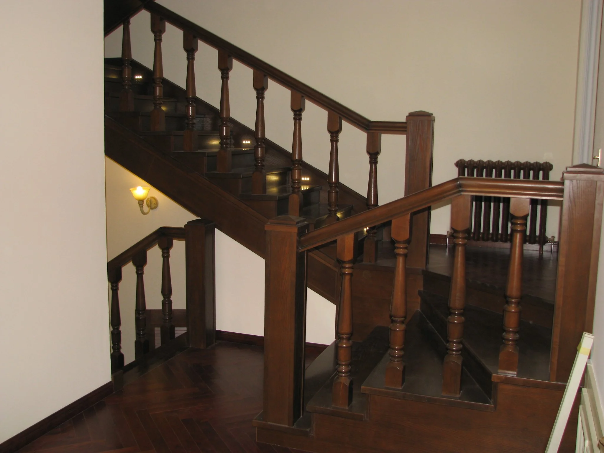 Wood staircase with ornate balustrades and handrails, interior lighting, beige walls, and a radiator