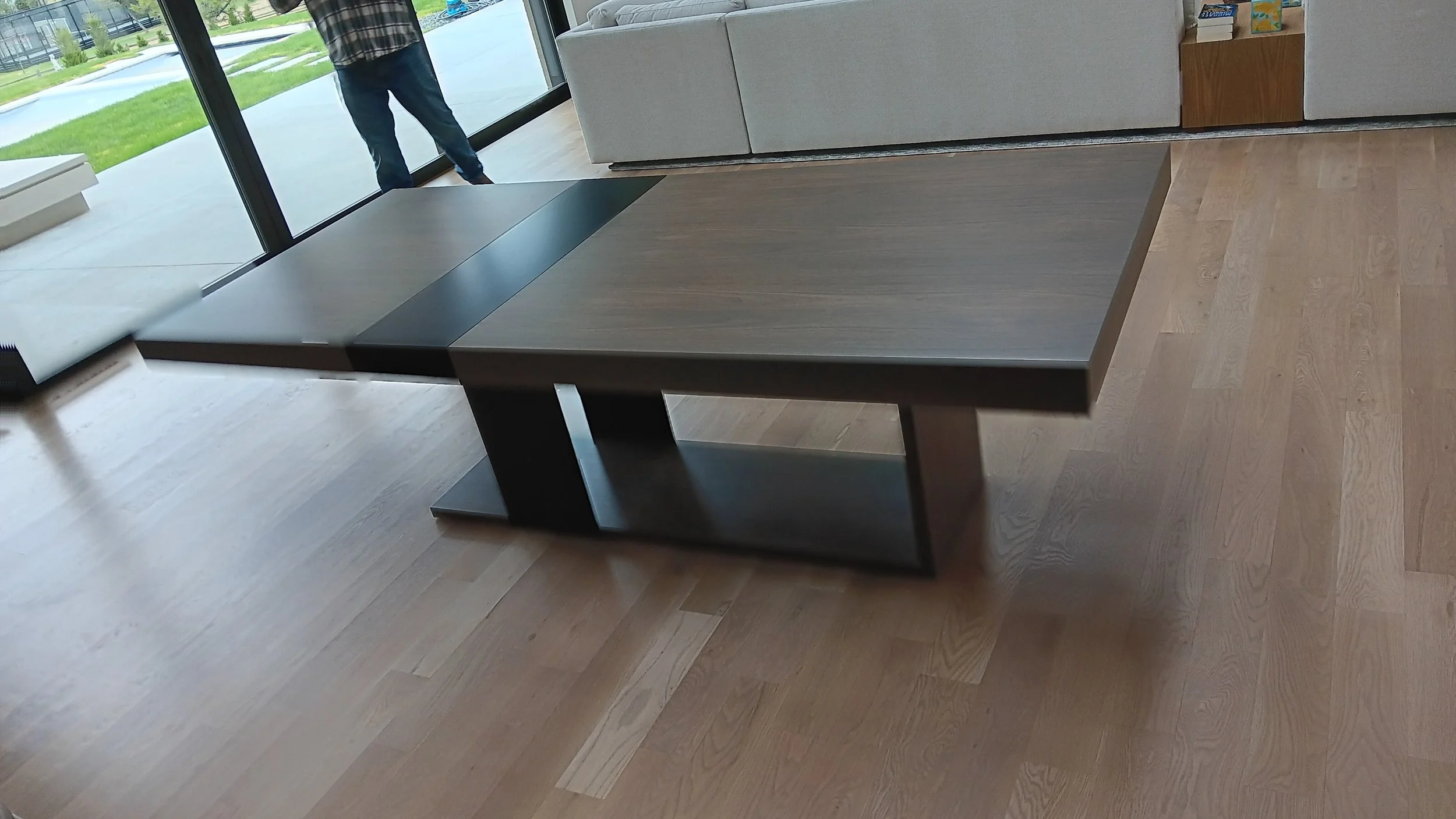 Modern black and wooden desk in a bright room with large glass windows, wooden floor, and a view of a green lawn outside.