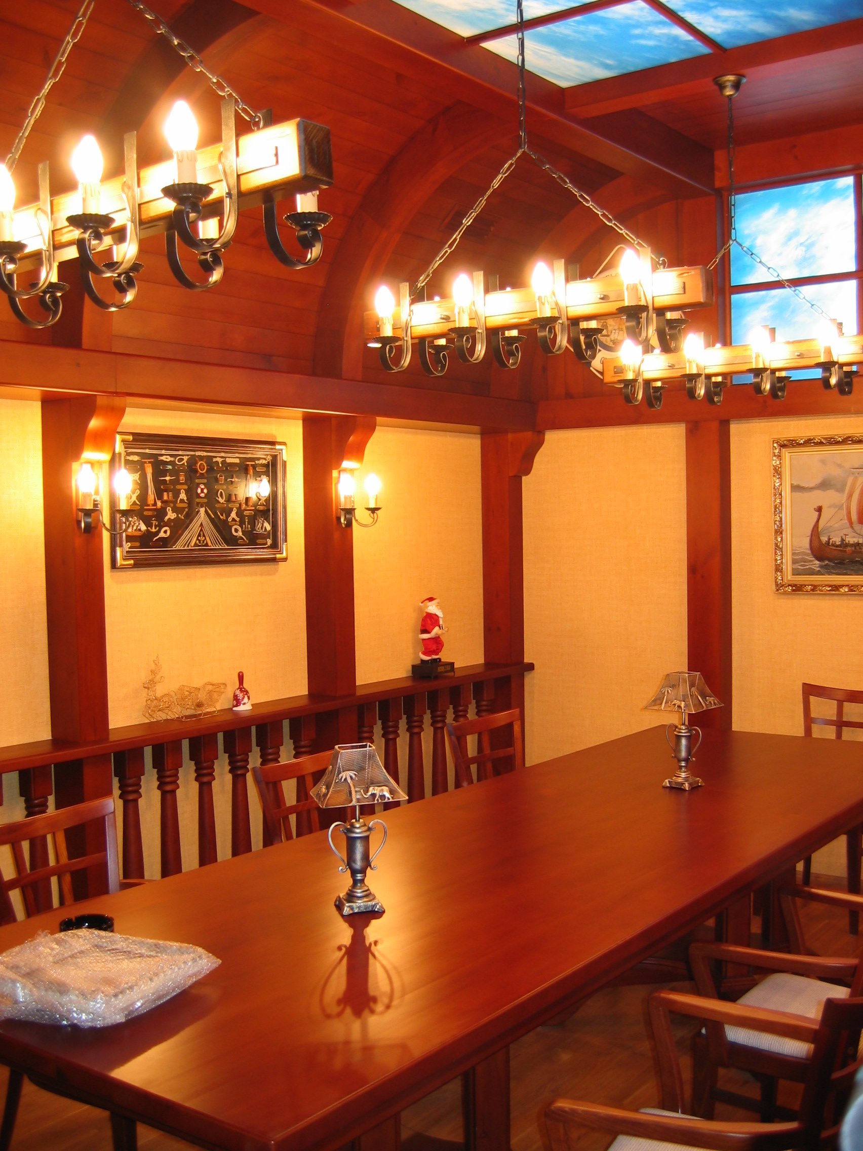 A dining room with a long wooden table and wooden chairs, decorated for Christmas with small lamps, festive figurines, and holiday-themed pictures on the walls, illuminated by chandelier and wall-mounted lights, with a ceiling that has a skylight sho