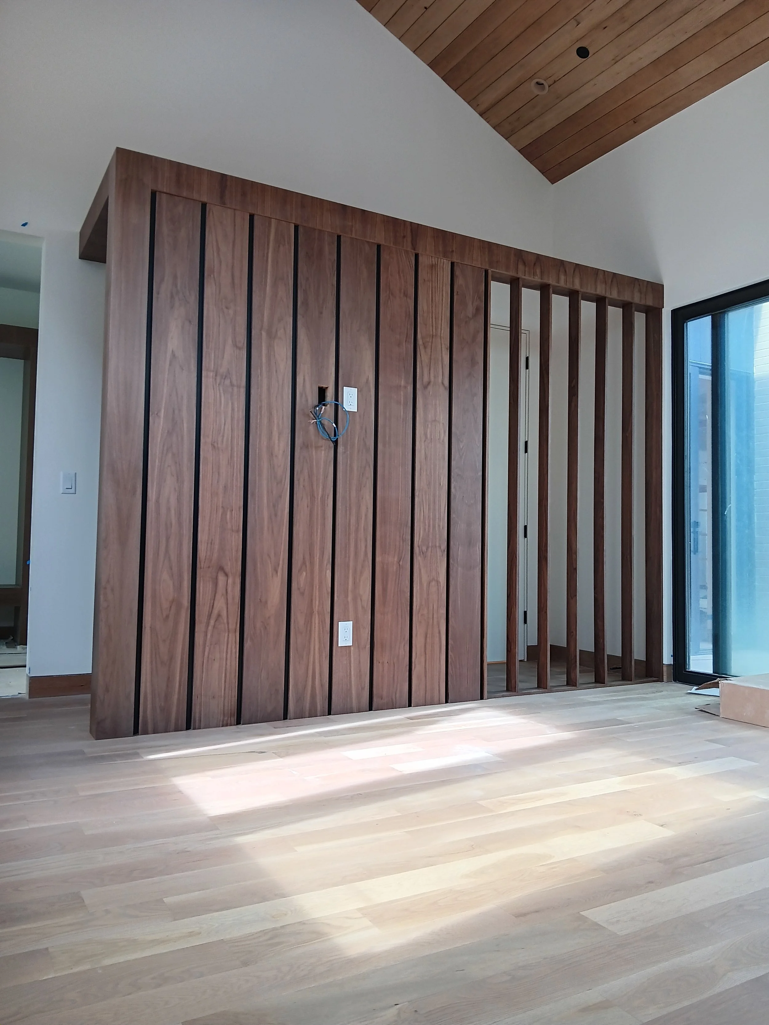 Interior of a modern room with wooden panel wall and vertical slats, hardwood flooring, and a sliding glass door letting in natural light.
