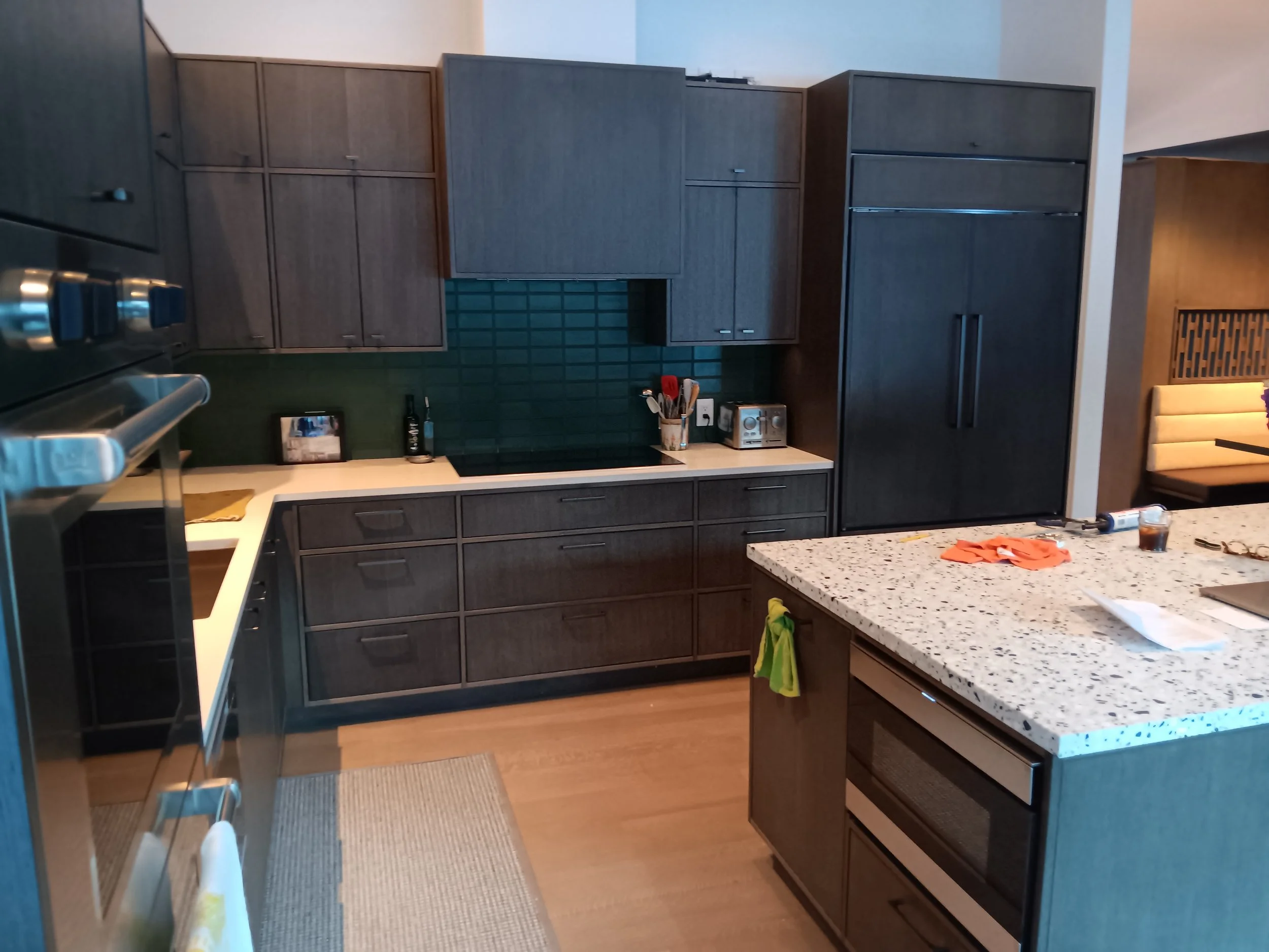 Modern kitchen with dark wood cabinets, green tiled backsplash, white countertop, and stainless steel appliances, including oven and toaster, with some items on the counter and a small island in the foreground.