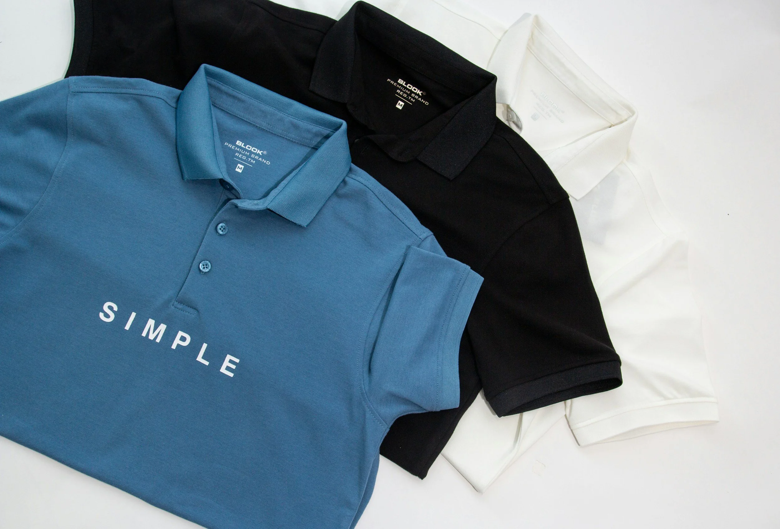 A flat lay of three cotton polo shirts in blue, black, and white, with the blue one prominently displaying the word 'SIMPLE' on its front.