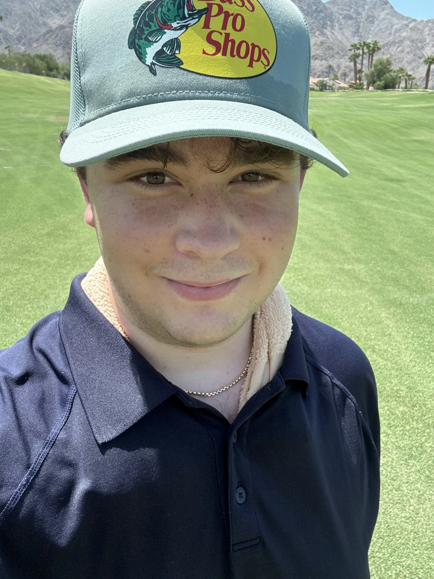 A young man taking a selfie on a golf course, wearing a gray cap with a Fish Pro Shops logo, a black collared shirt, and a gold chain necklace, with green grass, trees, and mountains in the background.