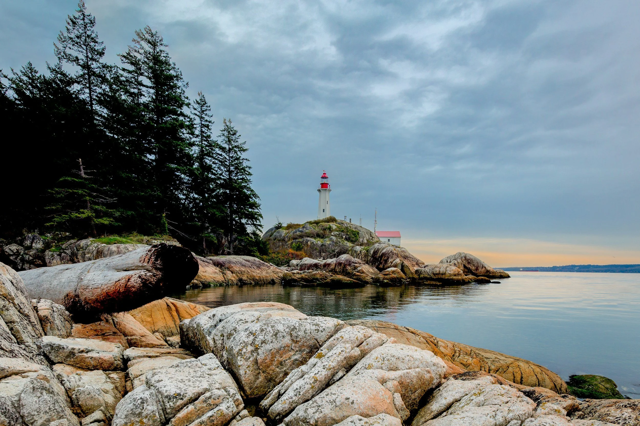A coastal landscape with large rocks in the foreground, a calm body of water, a lighthouse on a rocky hill, and trees on the left, under a cloudy sky.