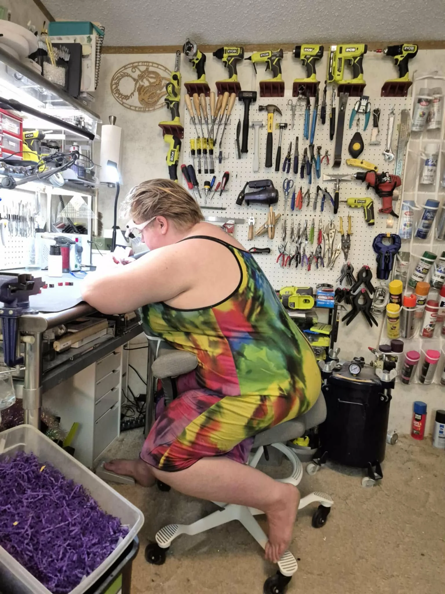 Chelsea Eldeen working at a cluttered workbench in her workshop, with tools hanging on a pegboard behind her and various supplies on shelves. She's wearing a colorful tie-dye dress and is barefoot, sitting on a Hag Capisco chair.