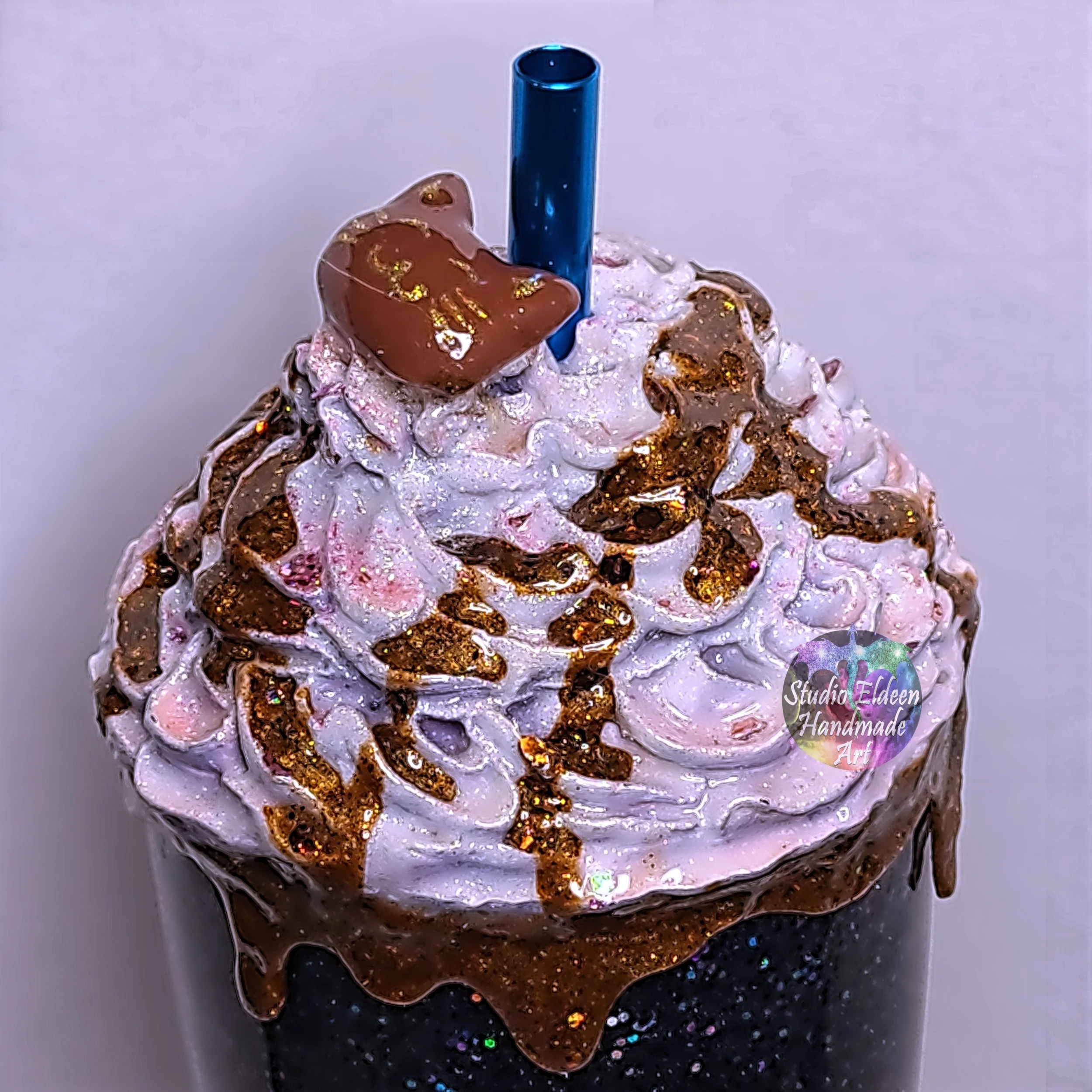 A milkshake with whipped cream, chocolate syrup, a cat-shaped chocolate piece, and a blue straw in a glittery black cup.