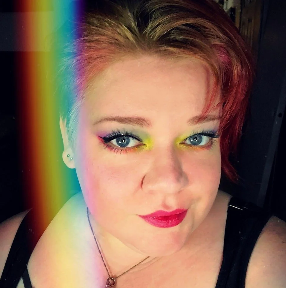 A woman with short red hair and blue eyes takes a selfie. She has colorful eye makeup, magenta lipstick, and wears a black top and a necklace. There is a rainbow-like light effect on the left side of the image.