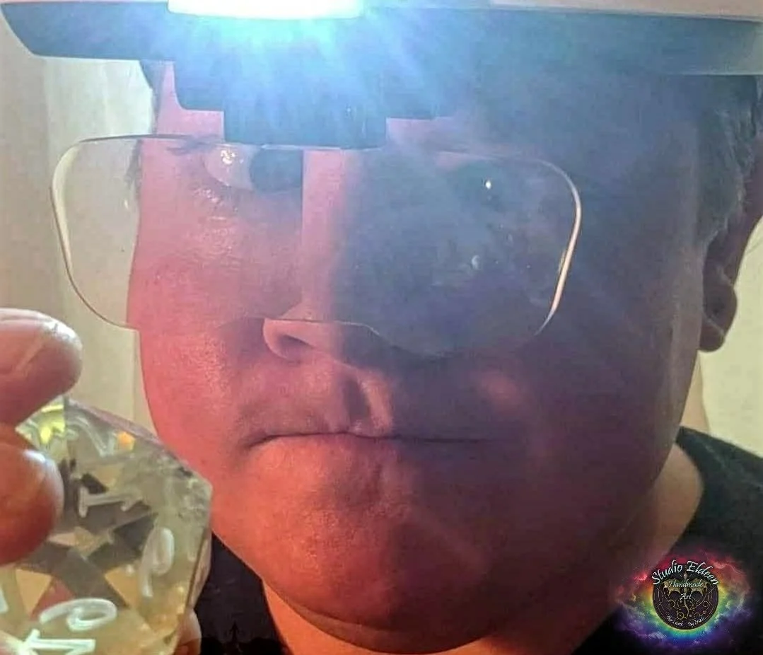 Chelsea Eldeen wearing lighted magnifying glasses holding a transparent d20. The magnifying glasses make her eyes look funny.
