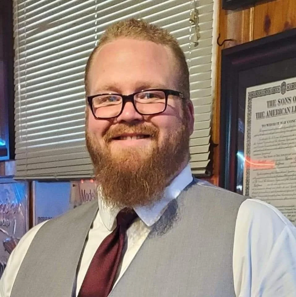 A smiling man with glasses, a beard, wearing a white shirt, tie, and grey vest, posing indoors near a window with blinds and framed certificate on the wall.