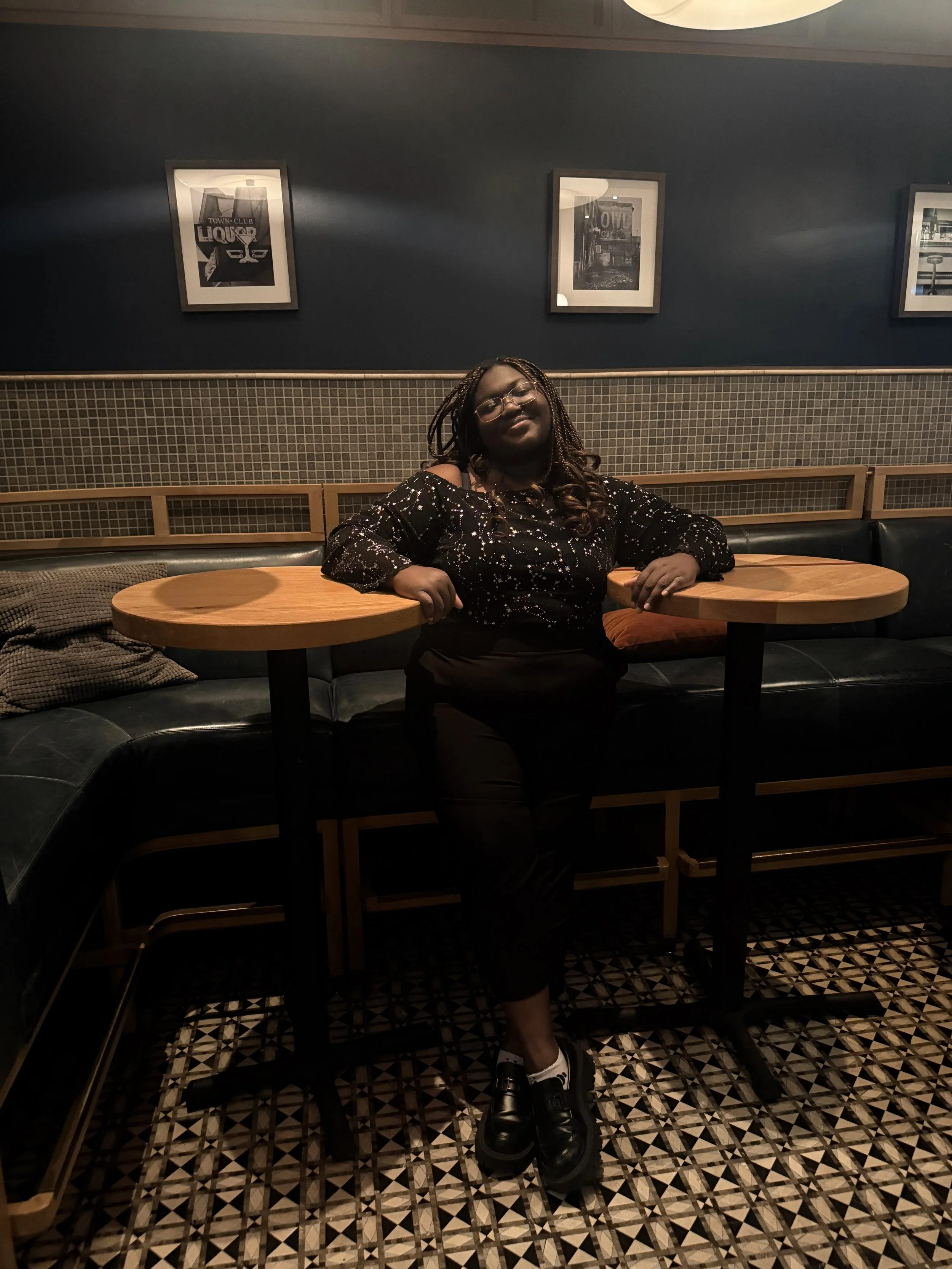 Young Black woman standing and smiling at the camera wearing a black shirt with constellations, and black pants and shoes. Arms are leaning on two tables.