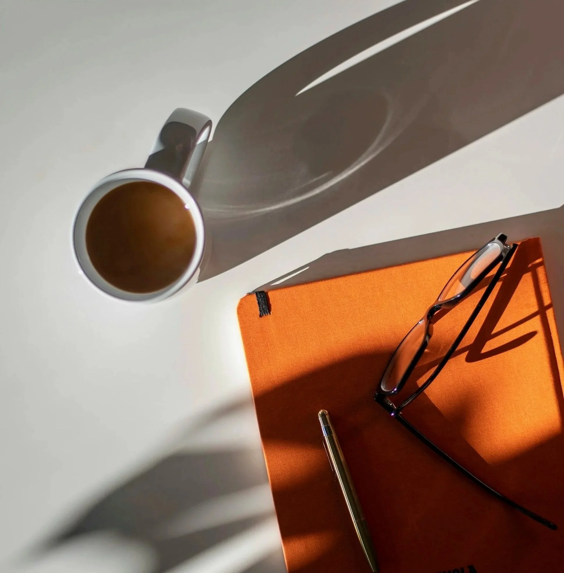 Top view of a white surface with a white mug filled with coffee, an orange notebook, a pair of black-rimmed eyeglasses, and a gold pen, with shadows cast by the objects.