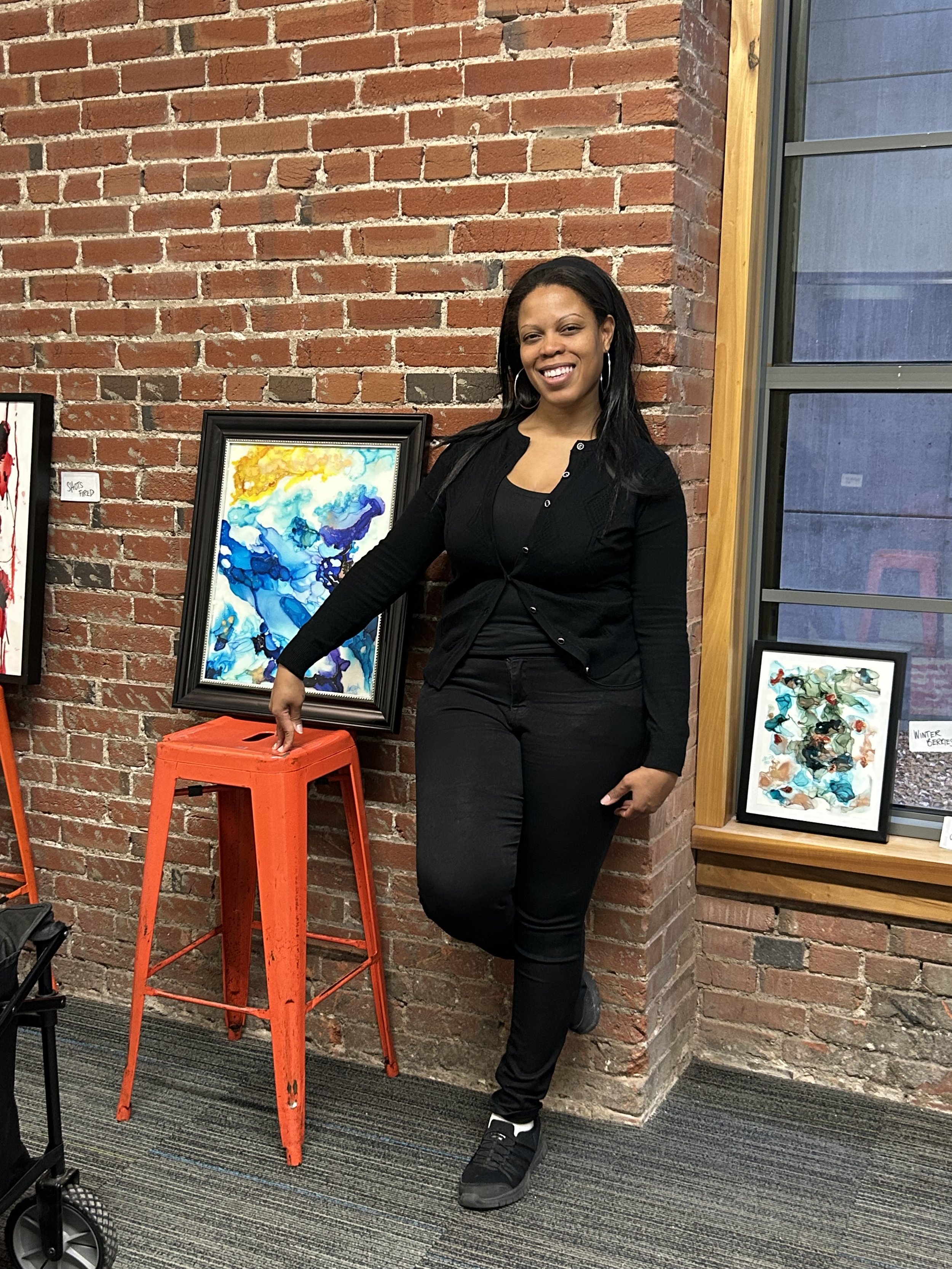 A woman with black hair and hoop earrings, dressed in black, standing against a brick wall and smiling. She has her left hand resting on a tall orange metal stool next to a colorful framed abstract painting. There are other artworks displayed nearby.