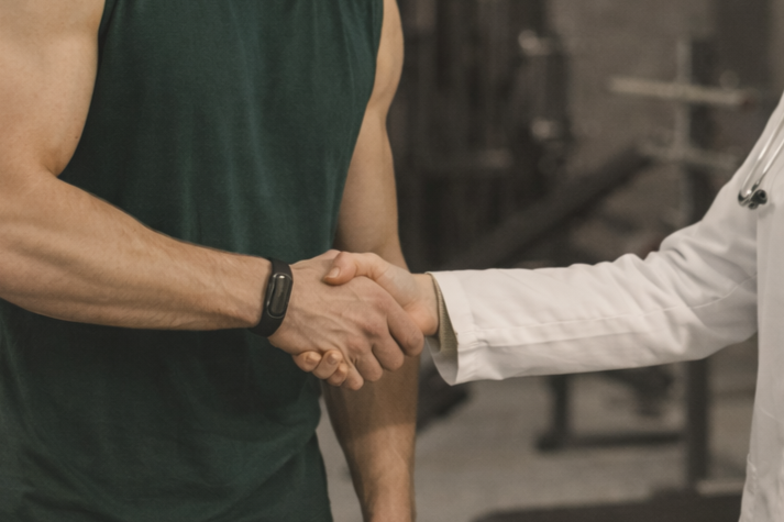 Close-up of a handshake between a man in a dark green shirt and a person in a white lab coat, in a medical or fitness setting.
