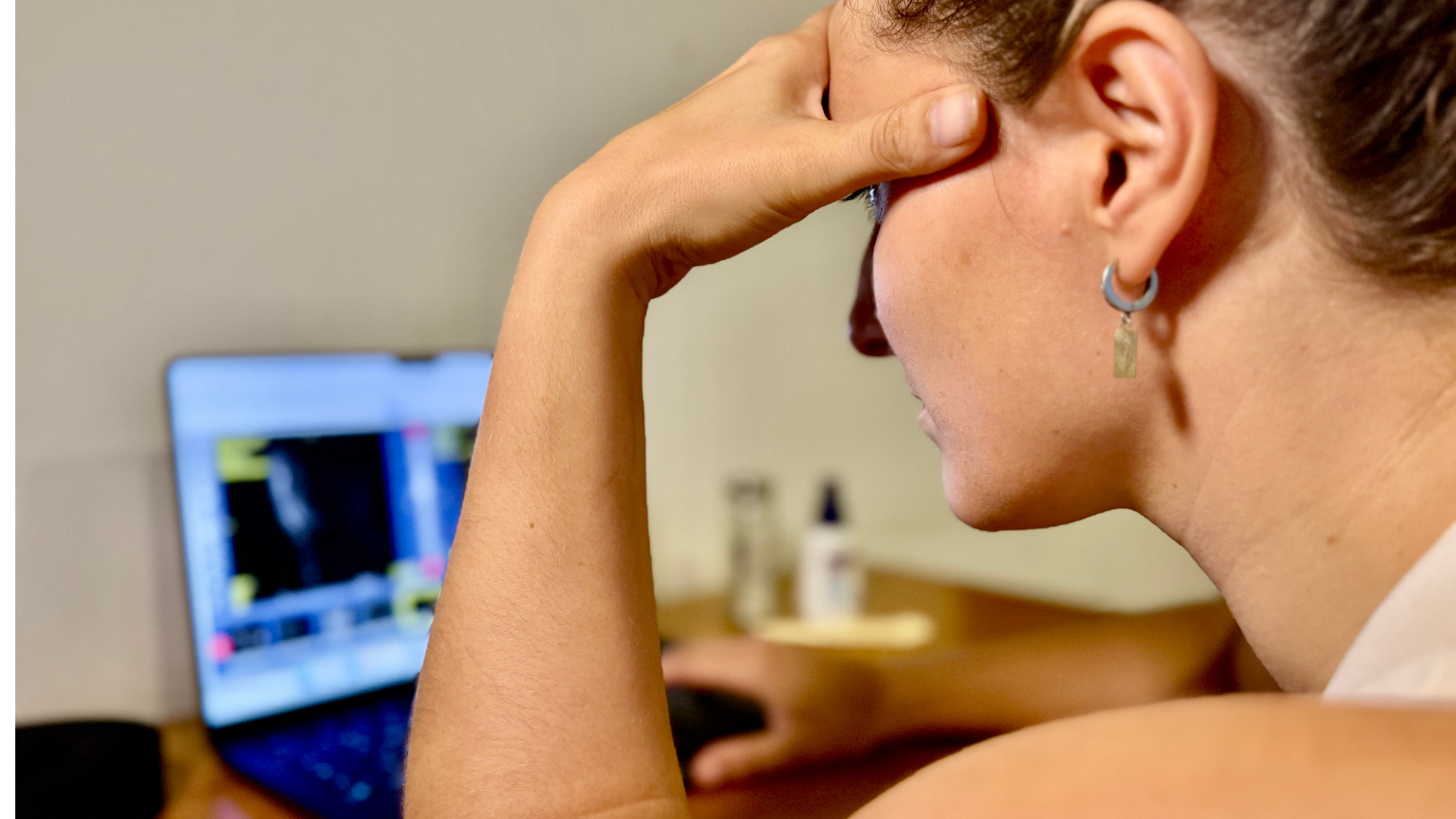 A woman sitting at a desk with her hand on her forehead, looking at a laptop screen displaying a blurred and colorful interface.