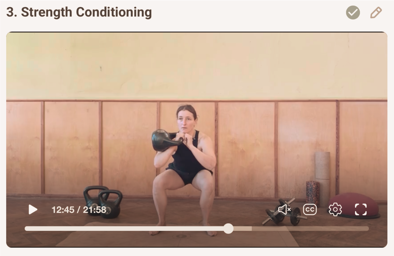 A woman in black workout attire squats while holding a kettlebell at chin level during a strength conditioning exercise in a gym or fitness studio. Various workout equipment, including a kettlebell, dumbbells, foam rollers, a exercise ball, and a yoga wheel, are placed on the floor around her.