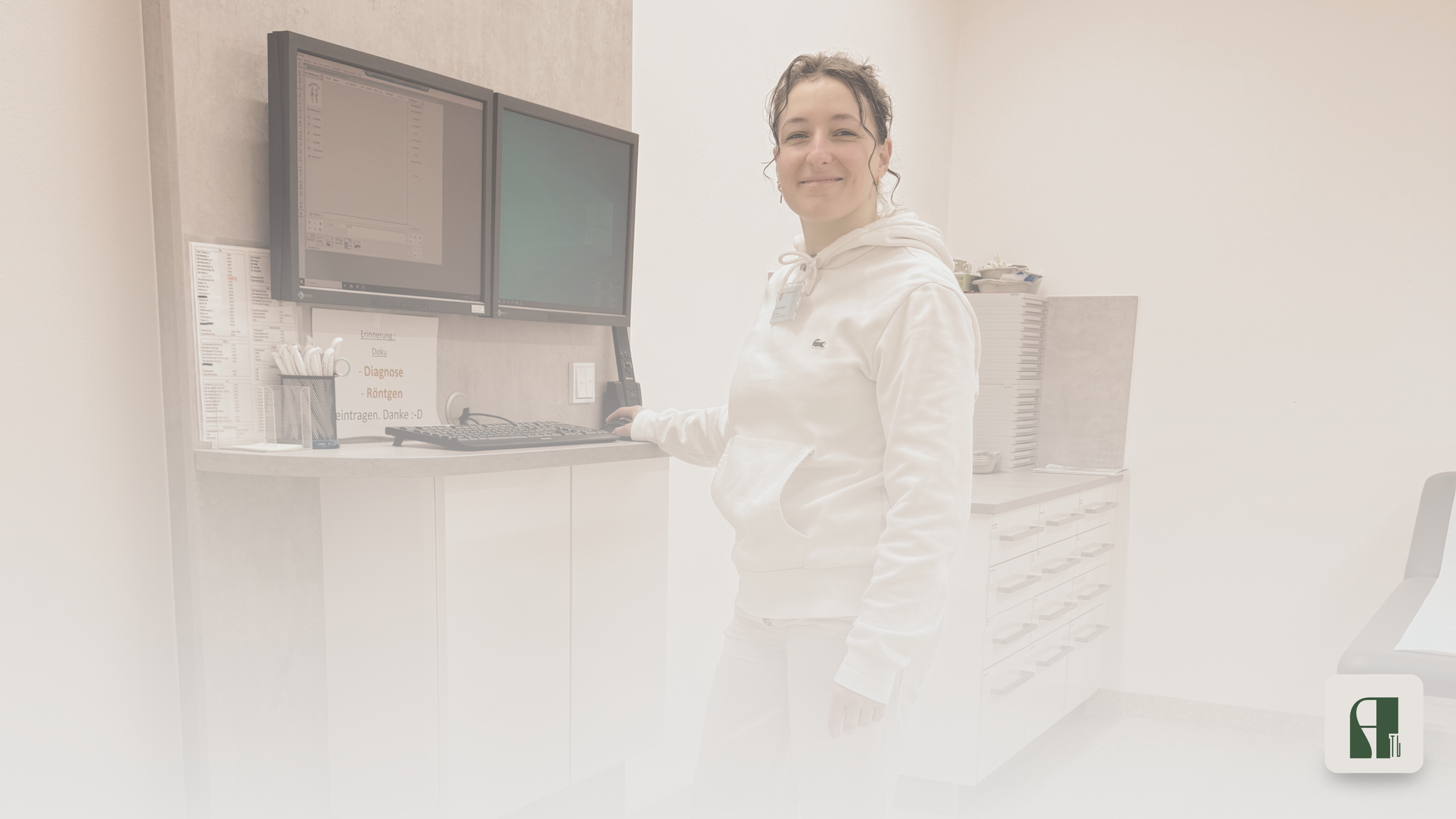 A woman in white hoodie standing at a computer workstation in a medical office, smiling at the camera.
