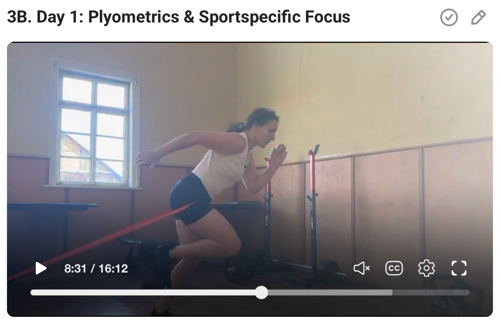 A woman in a white tank top and black shorts is performing a running exercise with resistance bands attached to her waist in a room with a window. 