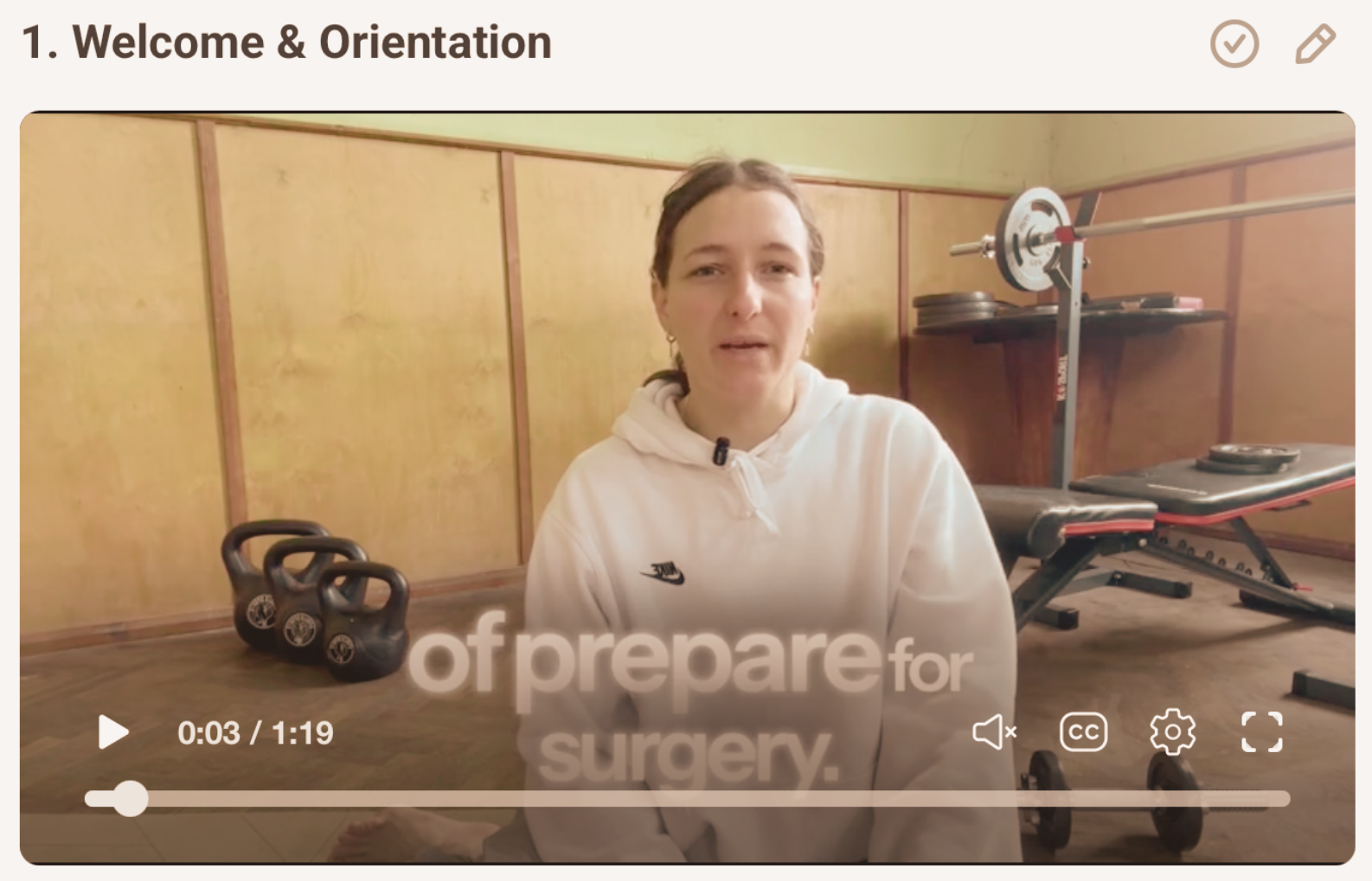 A woman in a white hoodie sitting in a gym or fitness room with weights and exercise equipment in the background, with partially visible text overlayed on the video saying 'of prepare for surgery'.